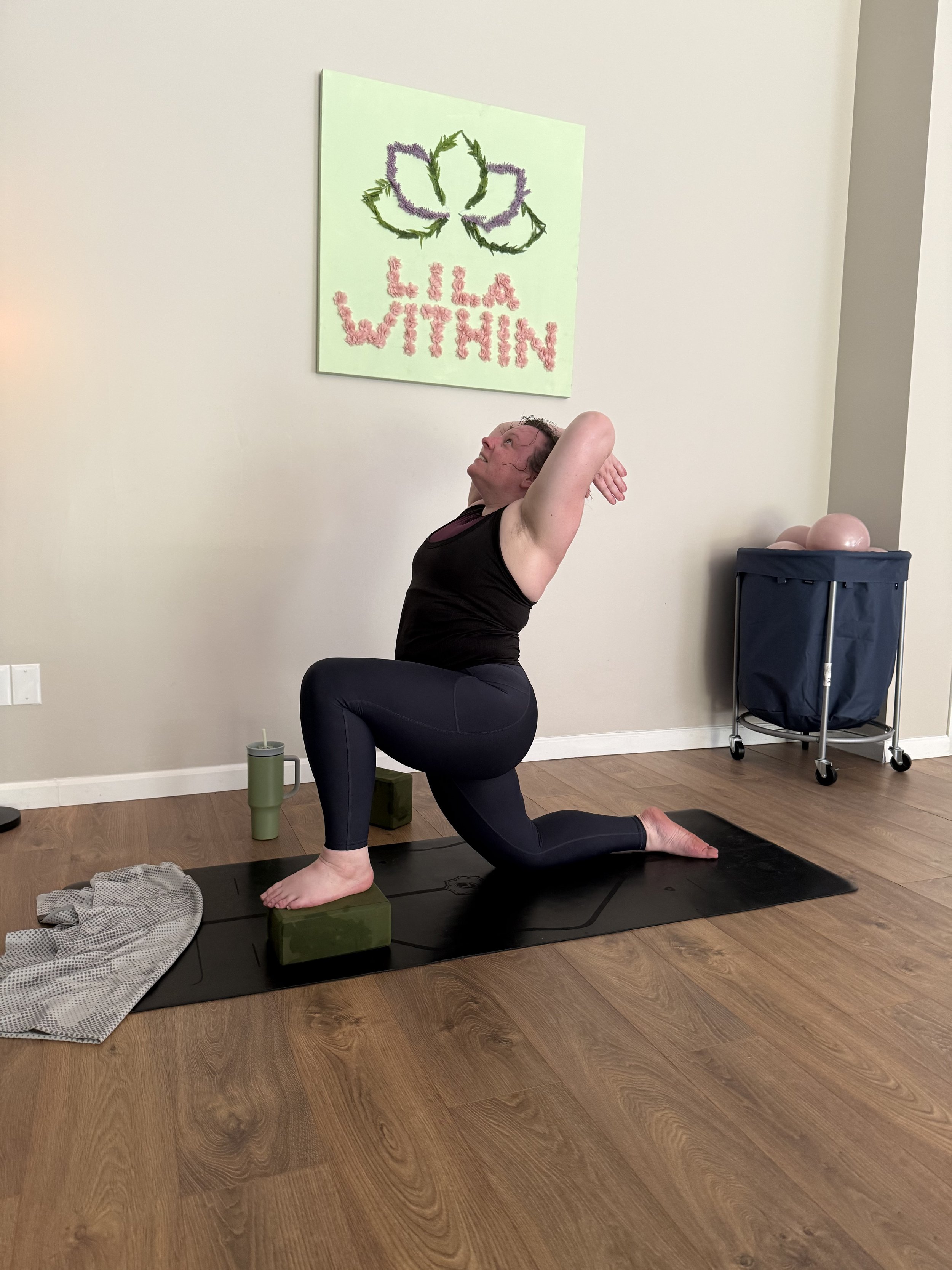 A woman practicing yoga in a lunge pose on a black yoga mat, with her hands behind her head, in a room with light-colored walls. A colorful sign behind her reads 'LILA WITHIN' with a flower drawing, and there are some exercise balls on a cart in the 