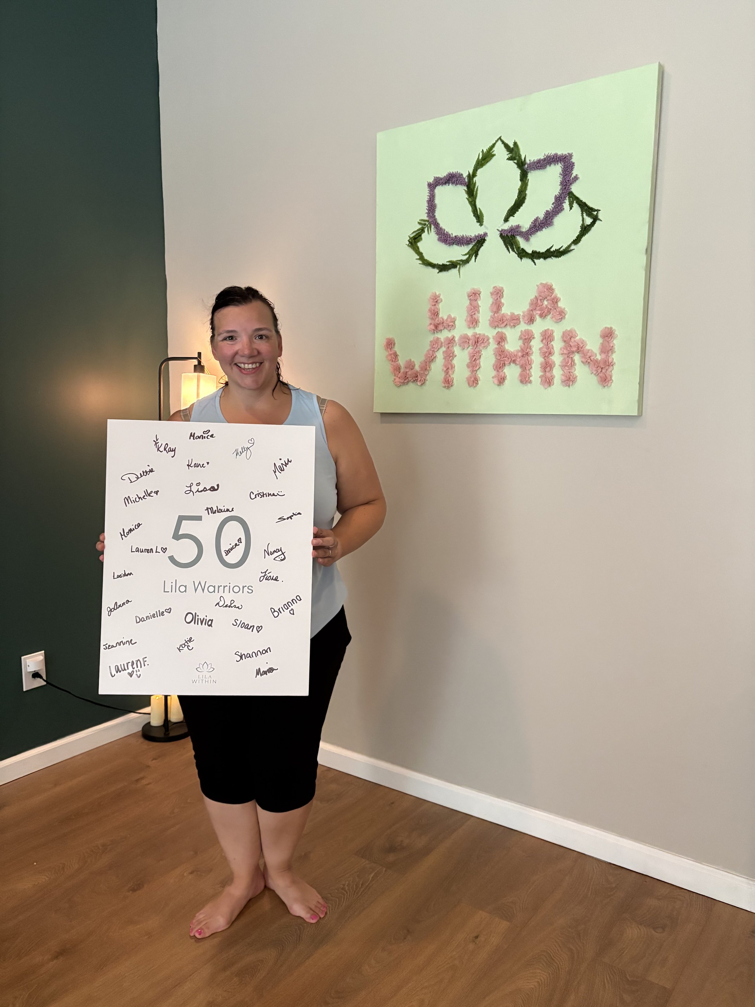 A woman standing on a wooden floor holding a large white sign with handwritten names around the number 50 and the words "Lila Warriors" at the bottom. She is barefoot, wearing a light blue sleeveless top and black shorts, smiling at the camera. Behin