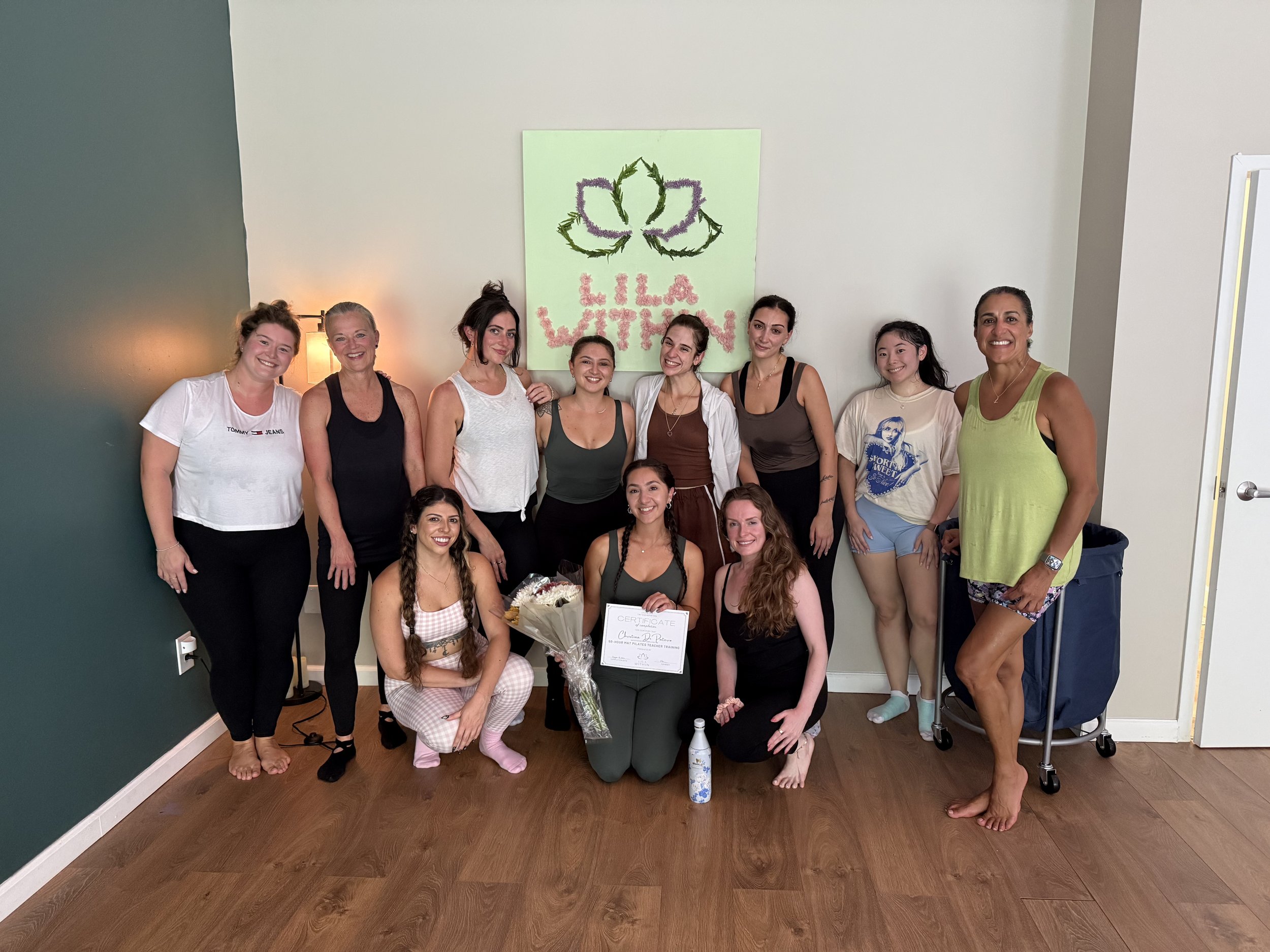 A group of ten women are posing together in a room. Some are standing, and some are kneeling or sitting. They are smiling and appear to be celebrating an achievement, as some are holding certificates and flowers. A large green poster with a floral de