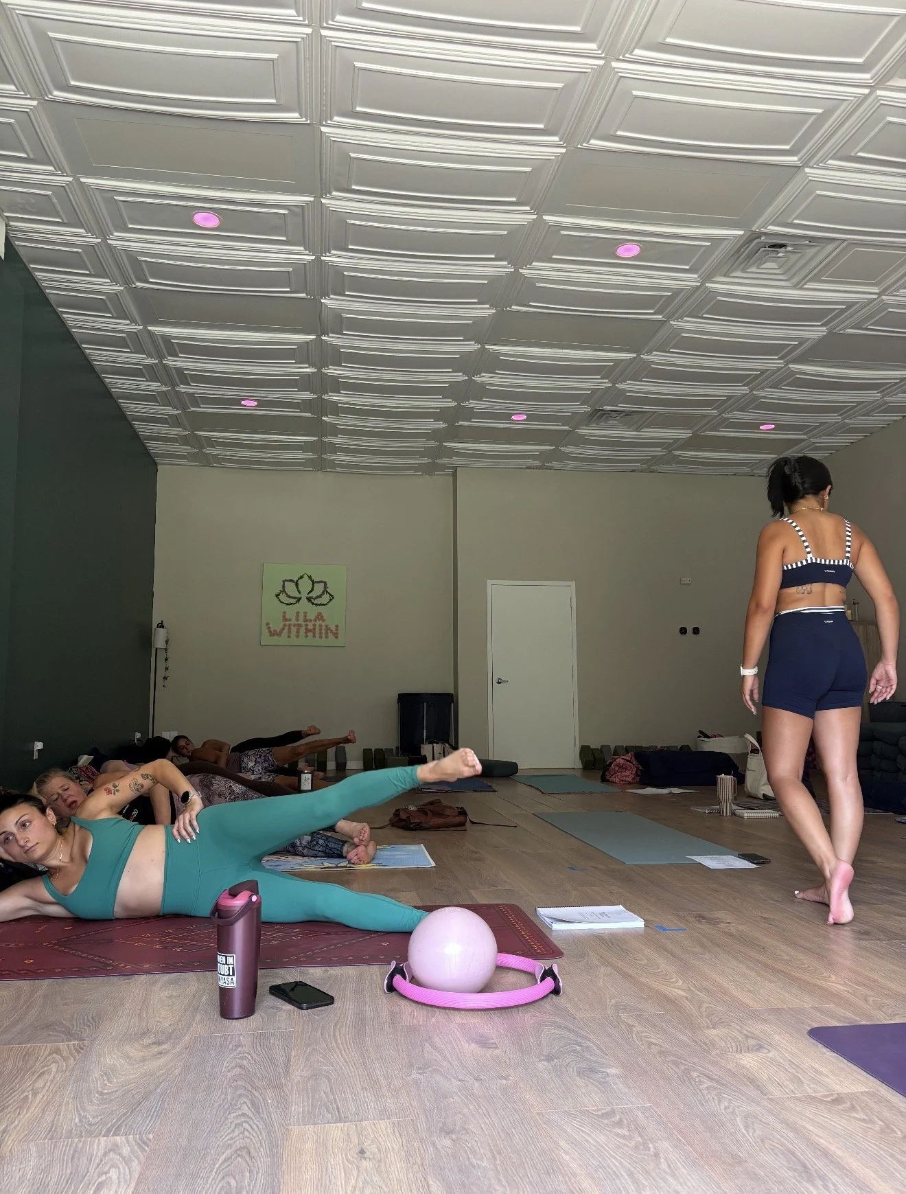 Women participating in a yoga or fitness class in a studio with ceiling panels and pink lighting. Some women are lying on mats, stretching or resting, while one woman stands at the front facing away from the camera. Equipment like a pink ball, water 