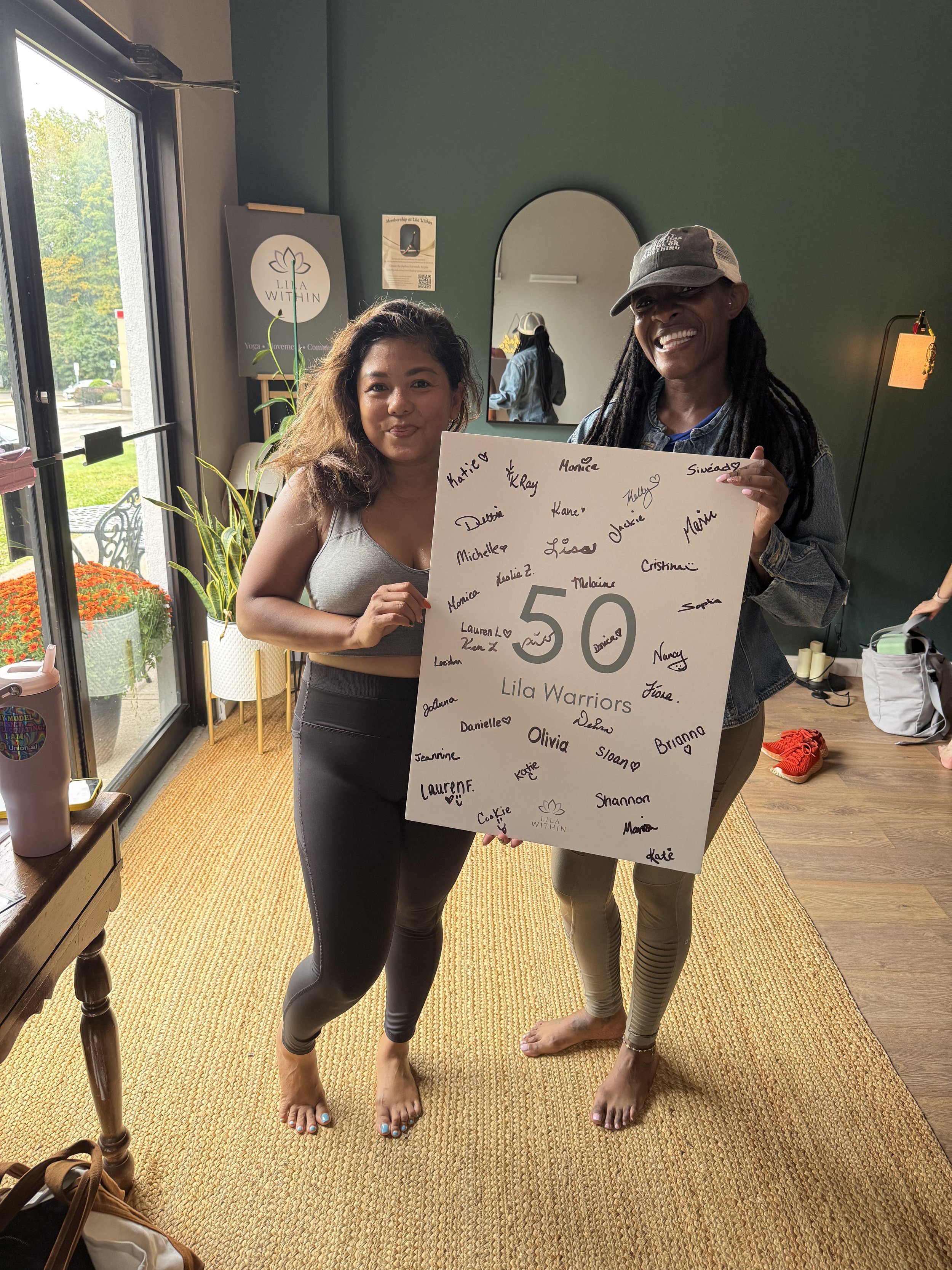 Two women standing barefoot indoors, holding a large white poster board with signatures and the text '50 Lila Warriors'. They are smiling, and behind them is a mirror and a window showing a green outdoor scene. The woman on the left is wearing a gray