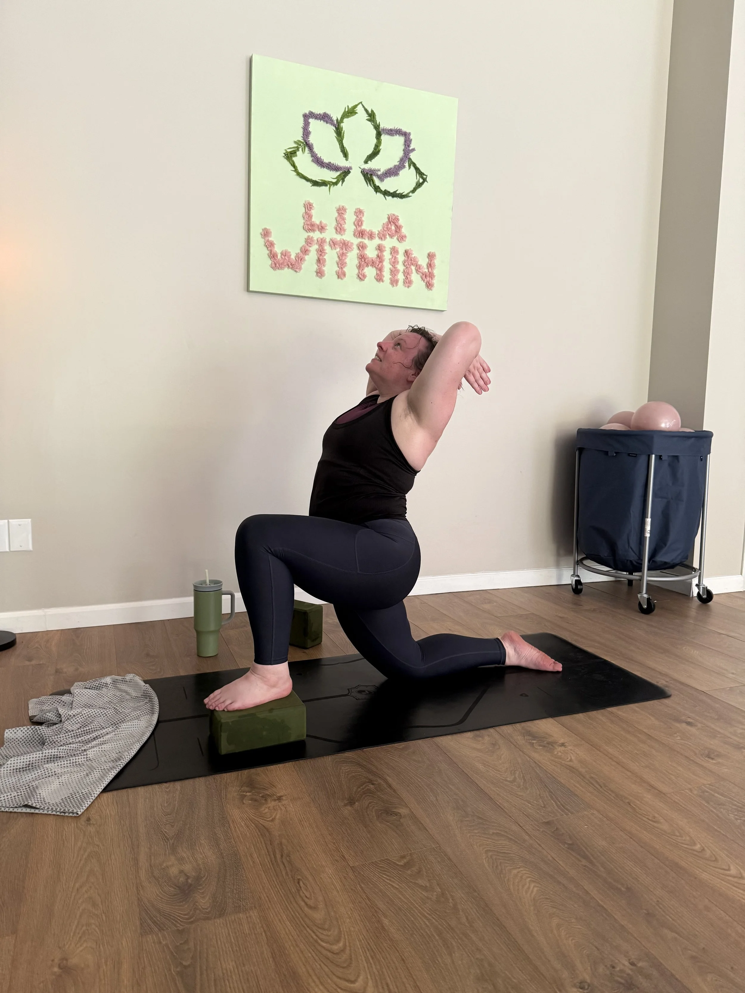 A woman practicing yoga indoors on a black mat, positioned in a low lunge pose with a slight backbend, using yoga blocks under her hands, with a canvass that has a green leaf and pink text on the wall behind her.