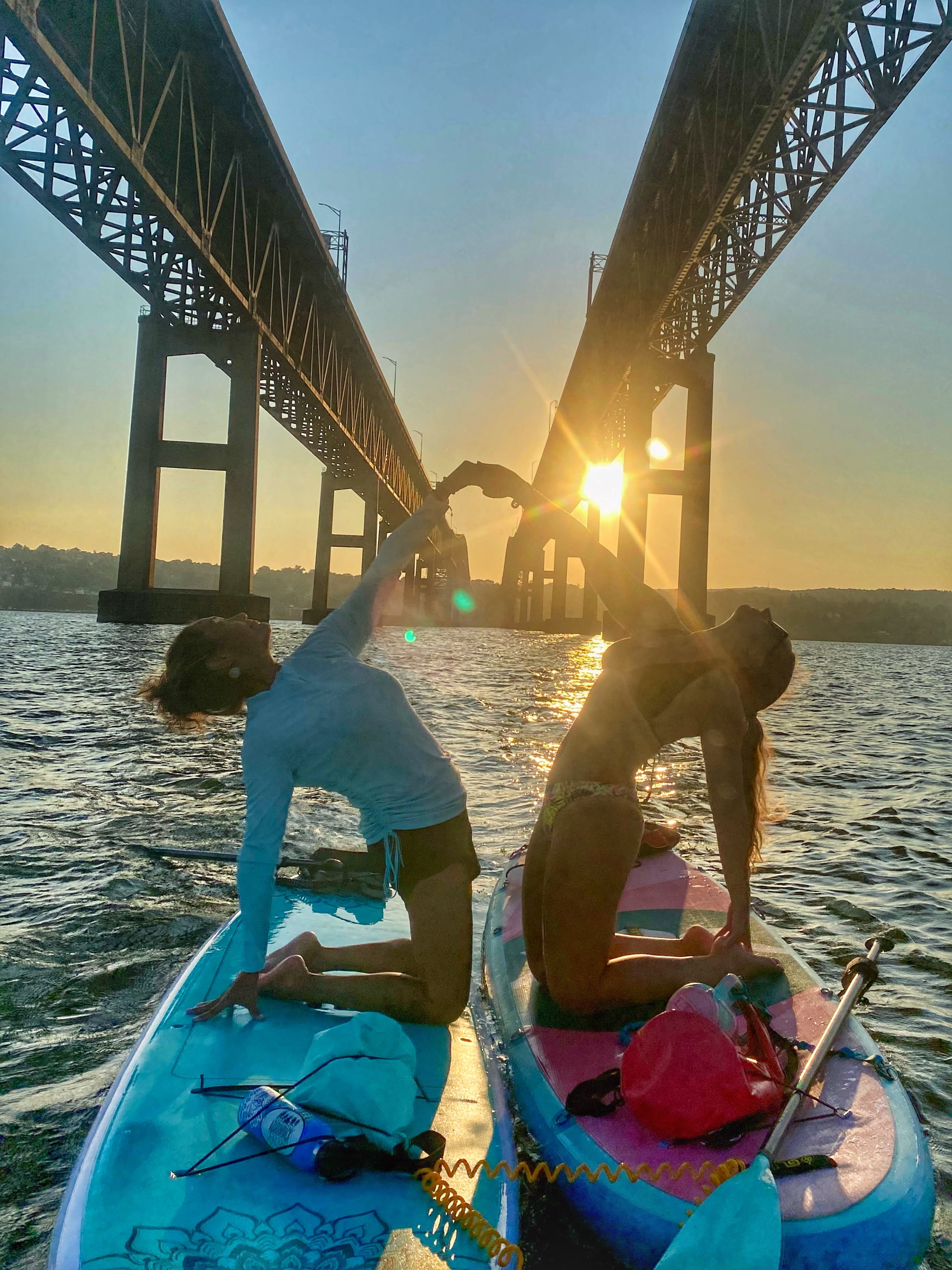 People practicing acro yoga on paddleboards during sunset on the water with a bridge in the background.