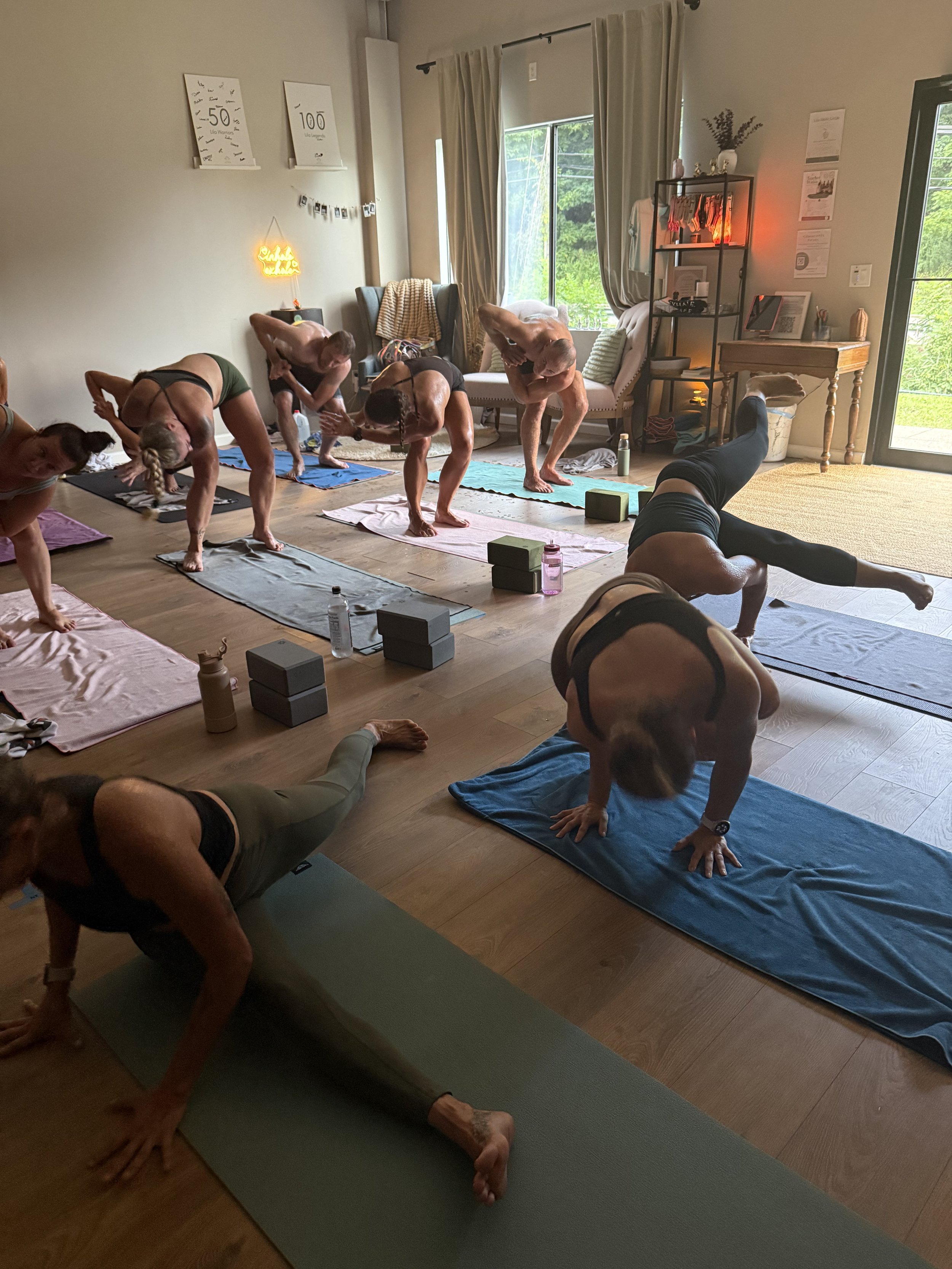 A group of people participating in a yoga class indoors, practicing downward dog pose on various colorful yoga mats. The room has large windows with curtains, a wooden floor, and shelves with decor, including a warm orange light, and outside greenery