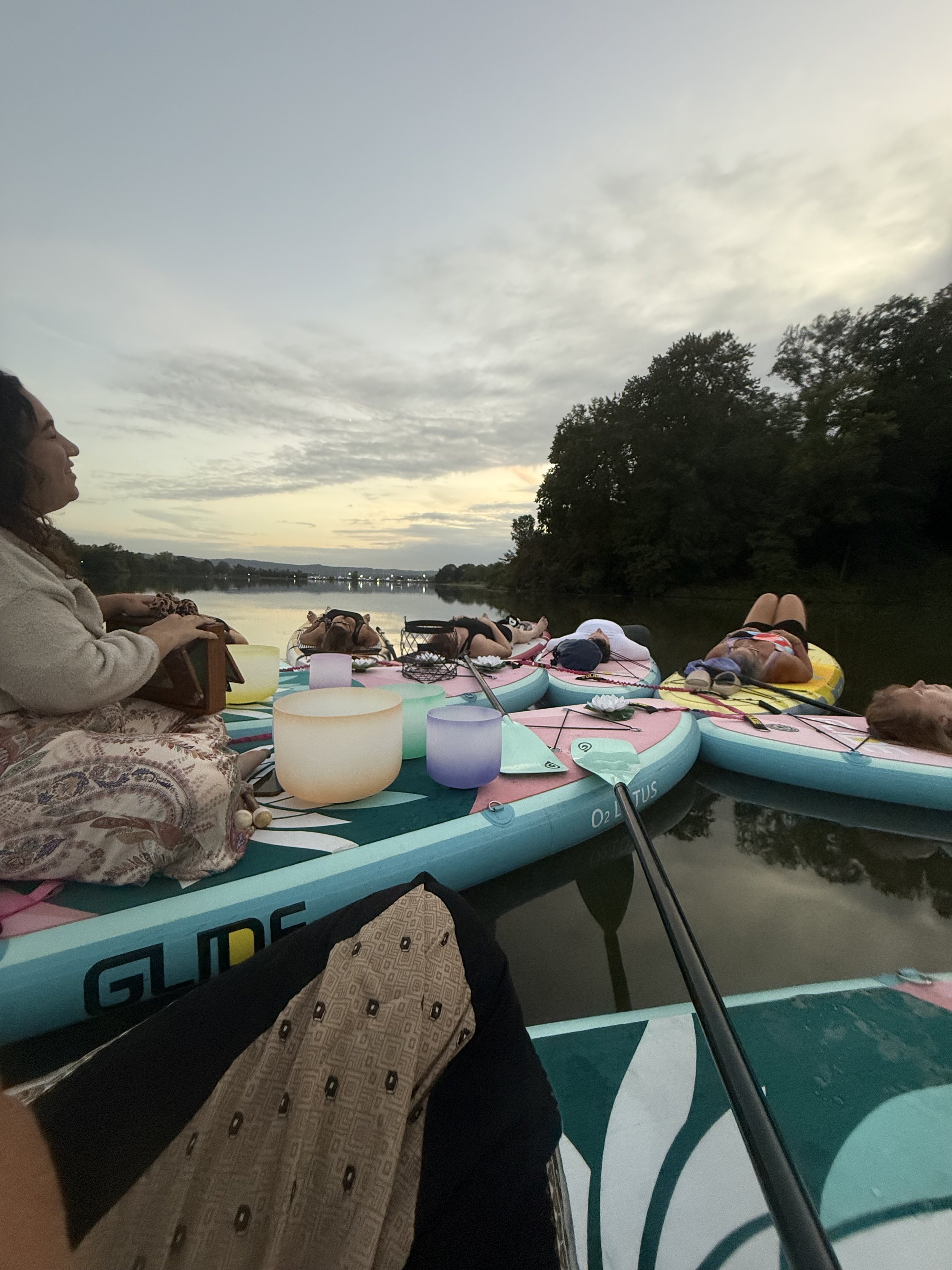 People relaxing on paddleboards by a river at sunset, with trees on the riverbank and a cloudy sky overhead.
