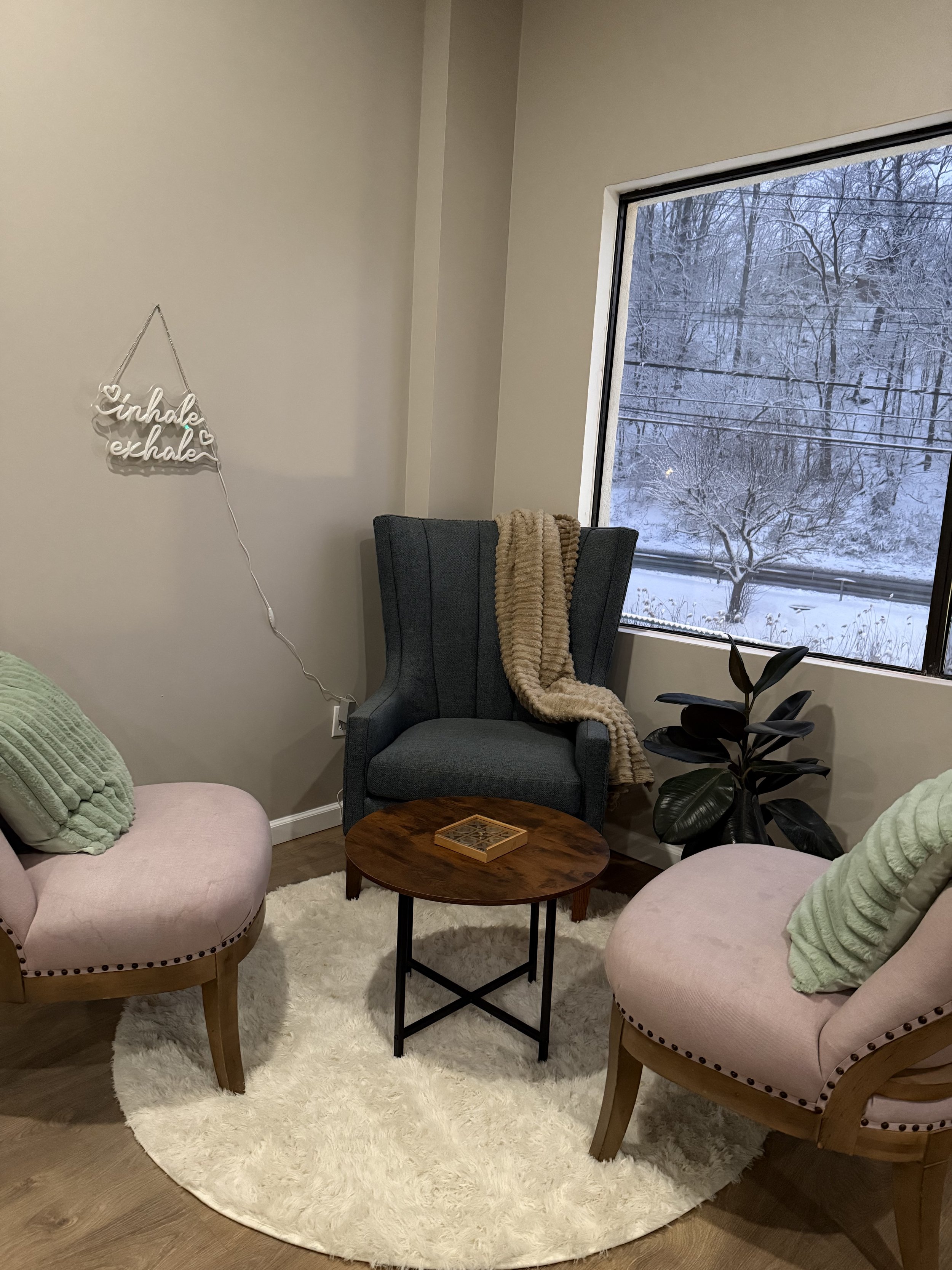 Living room with two pink chairs with green pillows, a middle wooden table, a dark gray armchair with a beige blanket, a large window showing snowy trees and ground outside, and a decorative illuminated sign on the wall reading 'inhale exhale'.
