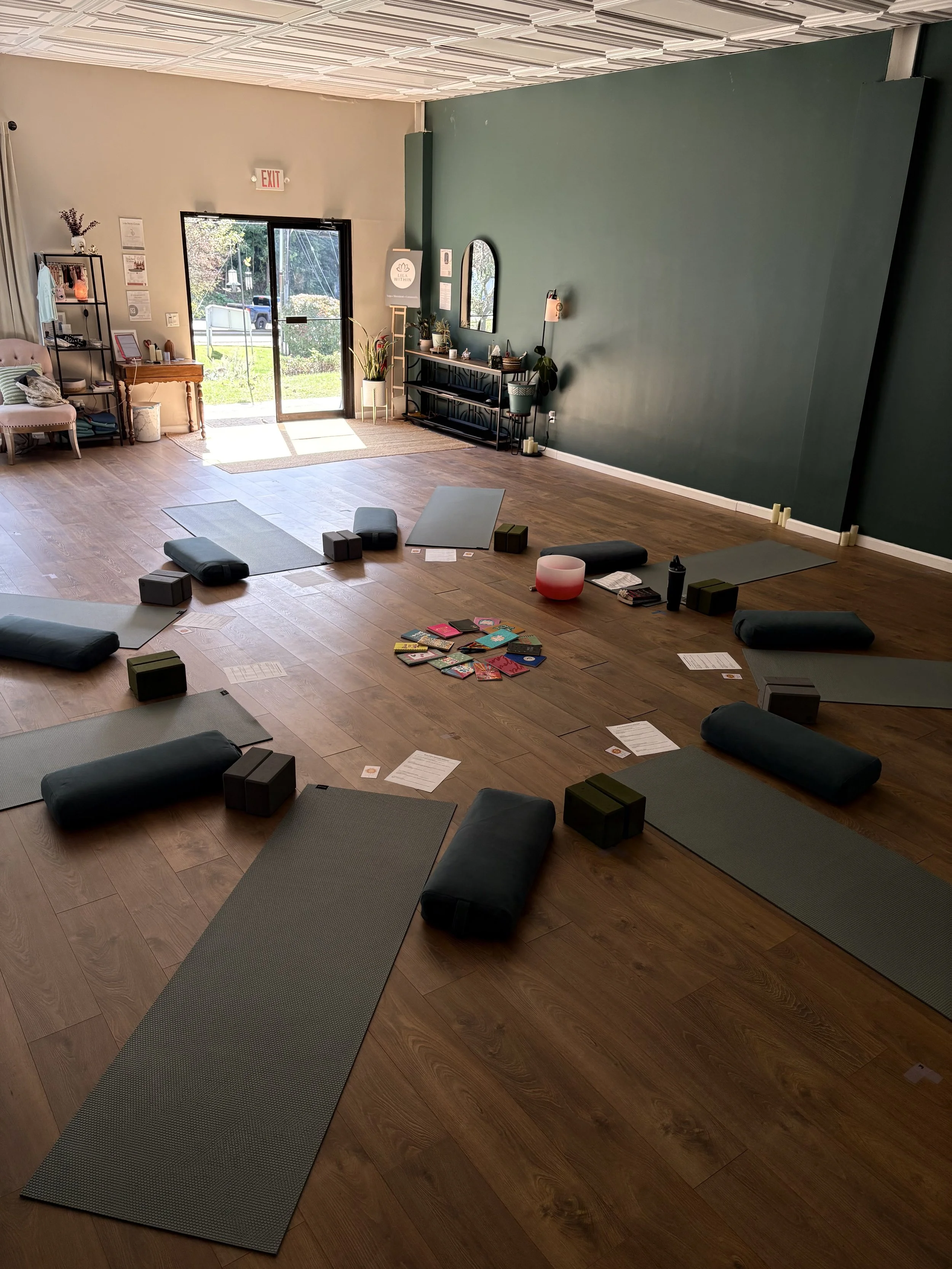 A yoga or meditation class setup with yoga mats, bolsters, and blocks arranged on a wooden floor inside a room with a green accent wall, large windows, and a glass door letting in natural light.
