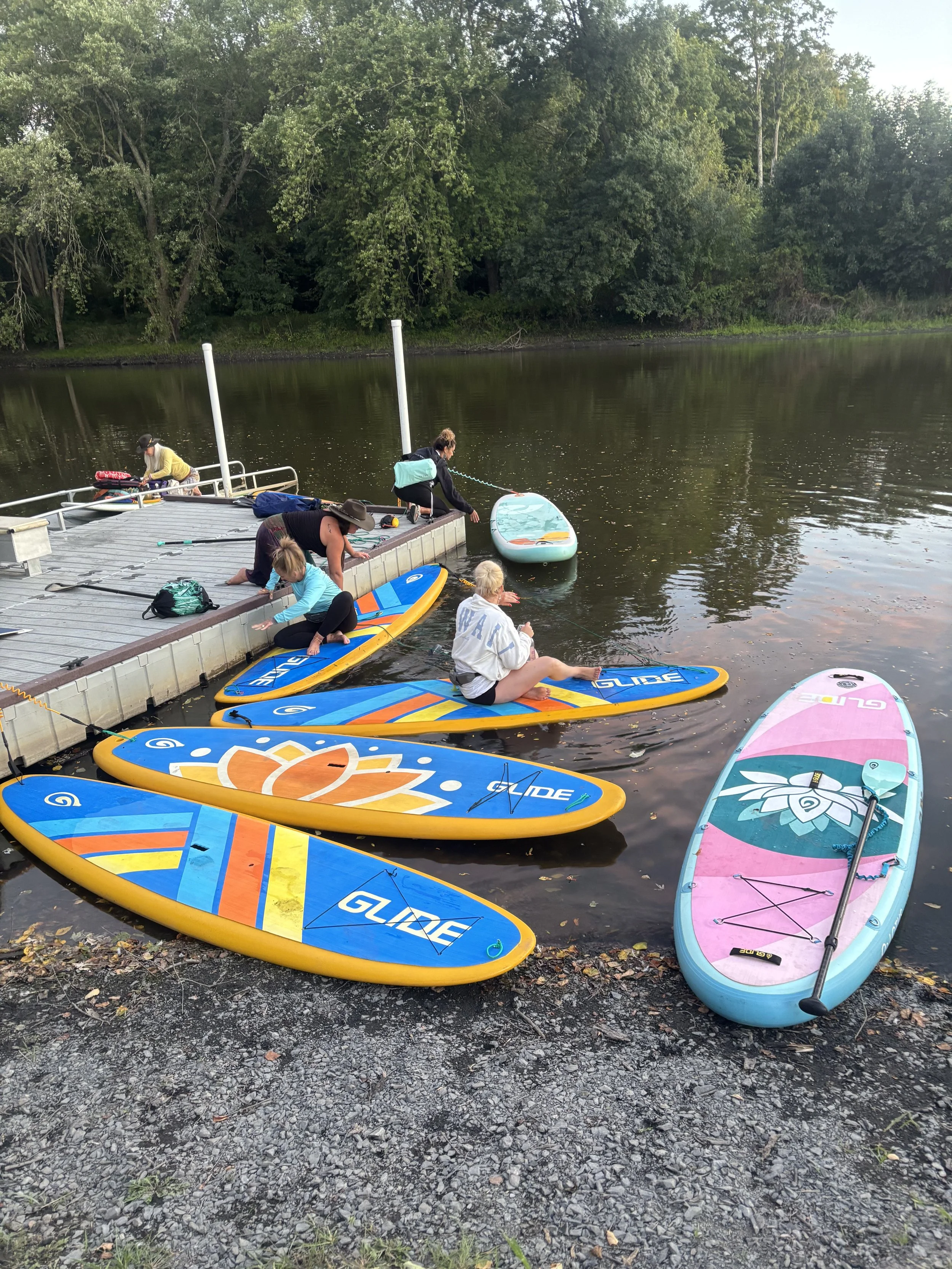 People preparing paddleboards on a dock by a river, surrounded by green trees.