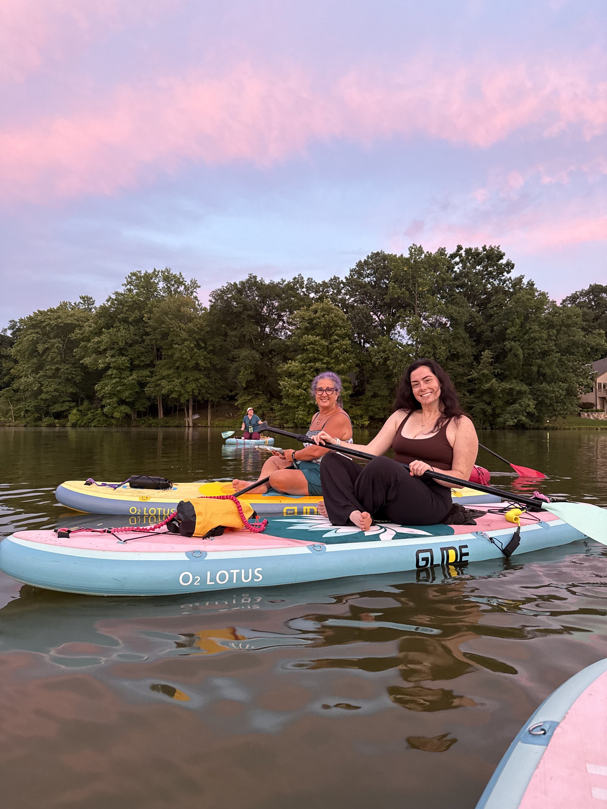 Two women sit on paddleboards on a calm body of water, smiling at the camera, with trees and houses visible in the background and a sky filled with pink and purple clouds during sunset.