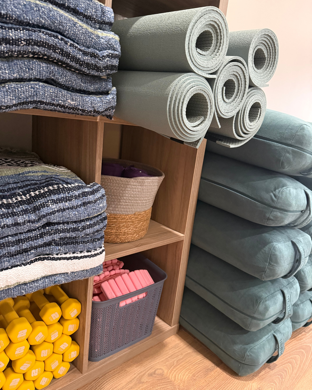 Stacked yoga mats, folded blankets, pink foam rollers, yellow dumbbells, and baskets on wooden shelves.
