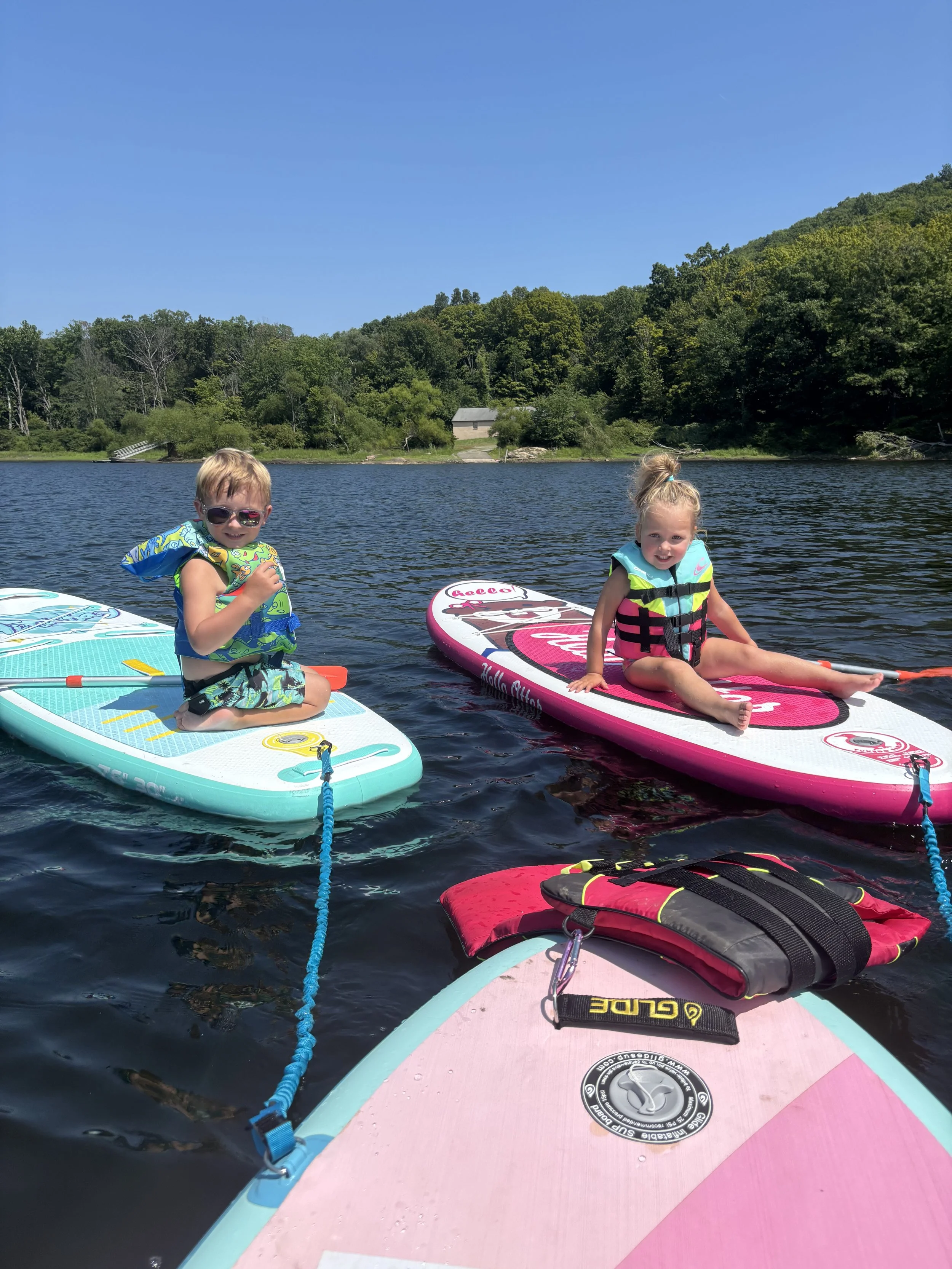 Two children sitting on paddleboards on a lake, with a lush green hillside and a small building in the background under a clear blue sky.