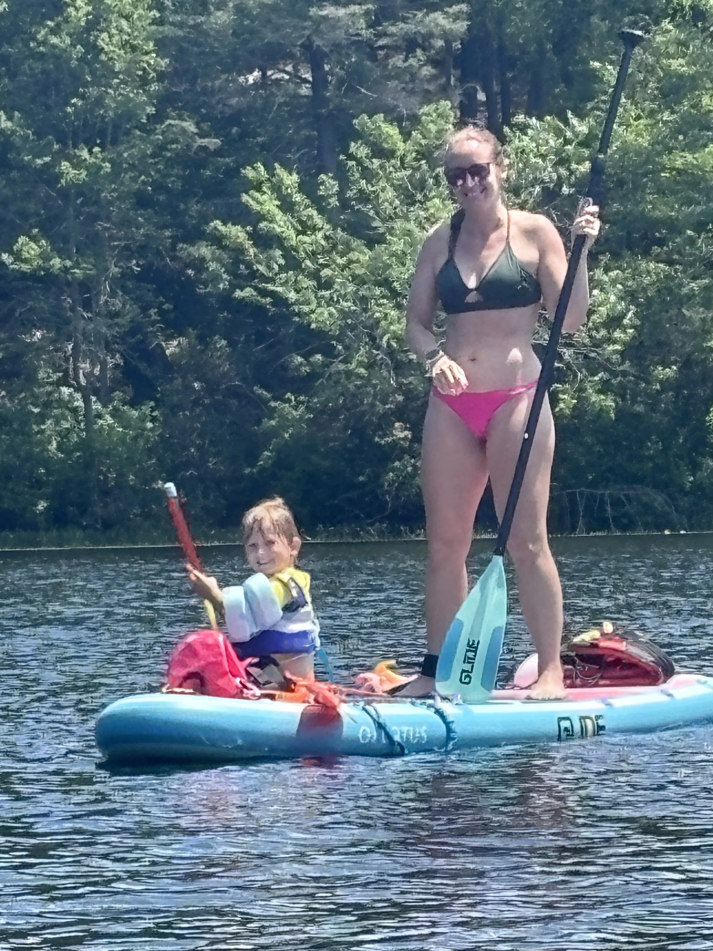 A woman in a bikini and sunglasses standing on an inflatable paddleboard, holding a paddle, on a lake with a forest in the background, smiling at the camera, with a child sitting on the paddleboard holding a toy.