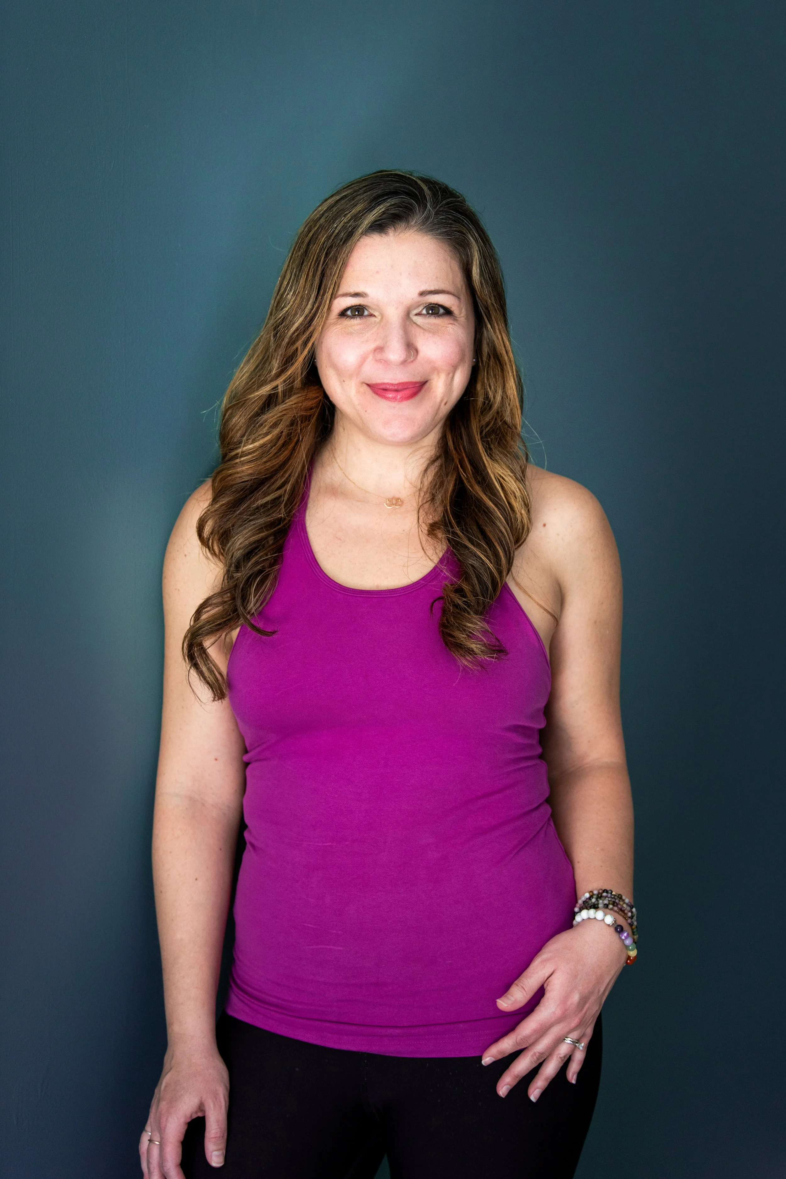 A woman with long, wavy brown hair wearing a sleeveless purple top, black pants, and multiple bracelets on her left wrist, standing against a dark blue wall and smiling at the camera.
