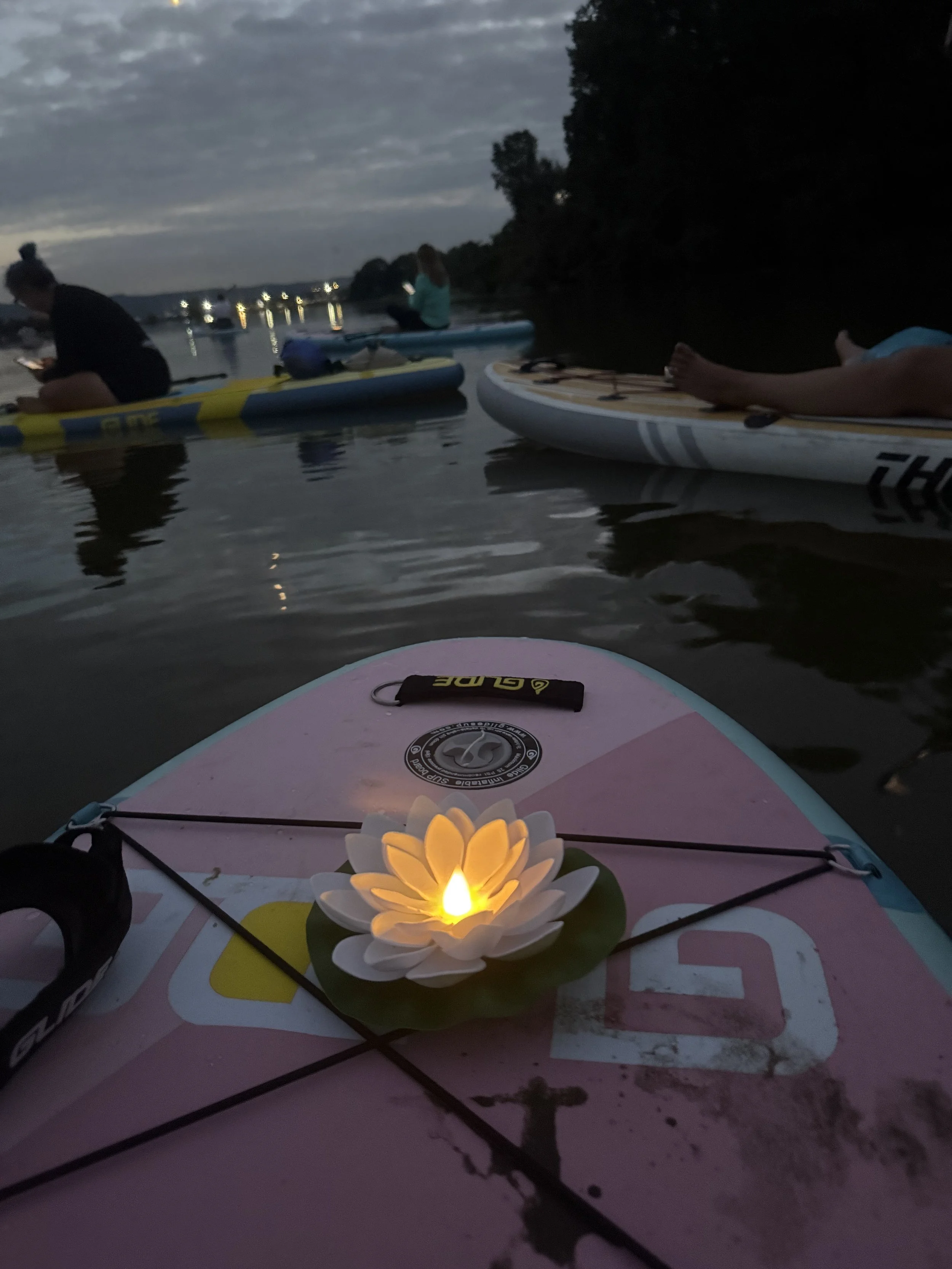 People paddleboarding on a lake at dusk with a decorative lit flower on the paddleboard in the foreground.