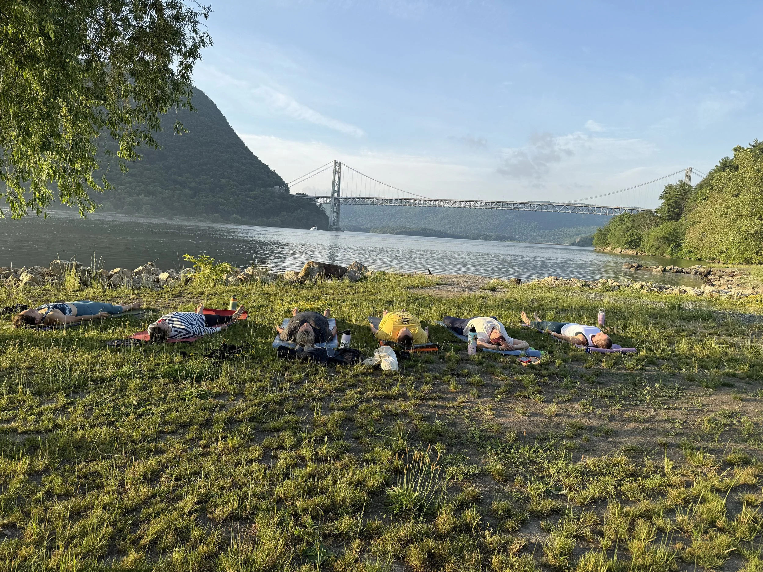 A group of people lying on their stomachs on yoga mats on a grassy riverside, with a large suspension bridge spanning across the river and green mountains in the background.