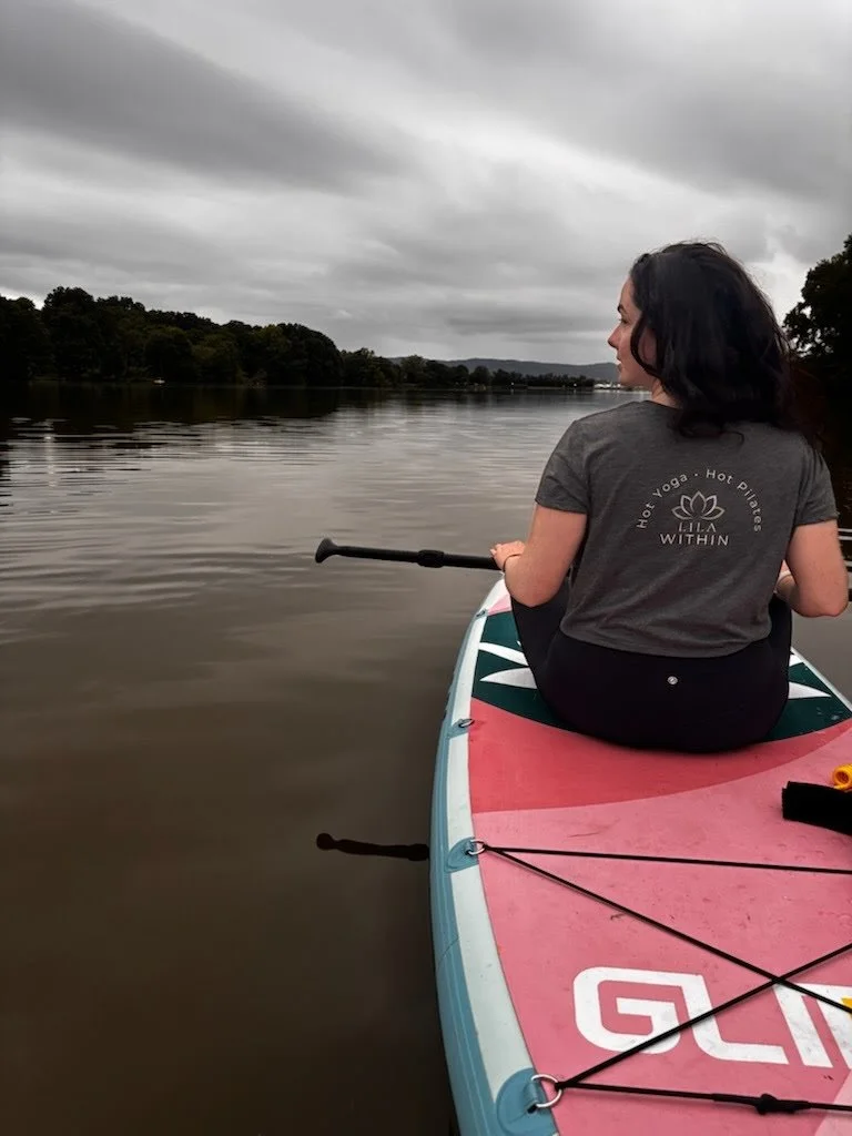 Woman sitting on a paddleboard on a calm lake, holding a paddle, with trees and cloudy sky in the background.