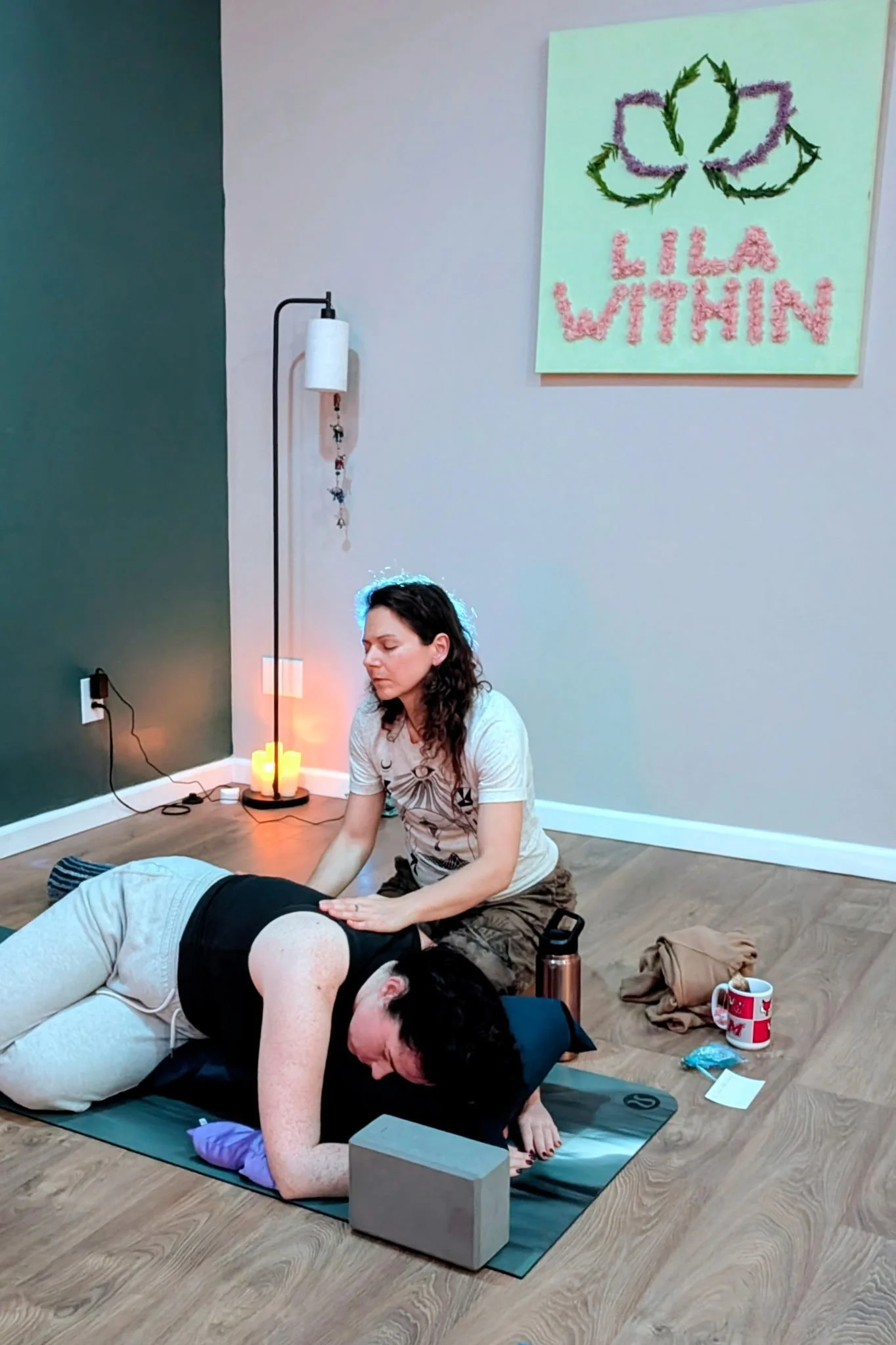A woman practicing yoga or receiving a massage while a person lies face down on a yoga mat in a cozy indoor setting with candles, a decorative sign that says 'Lila Within,' a tall floor lamp, and personal items like a mug and clothing on the floor.