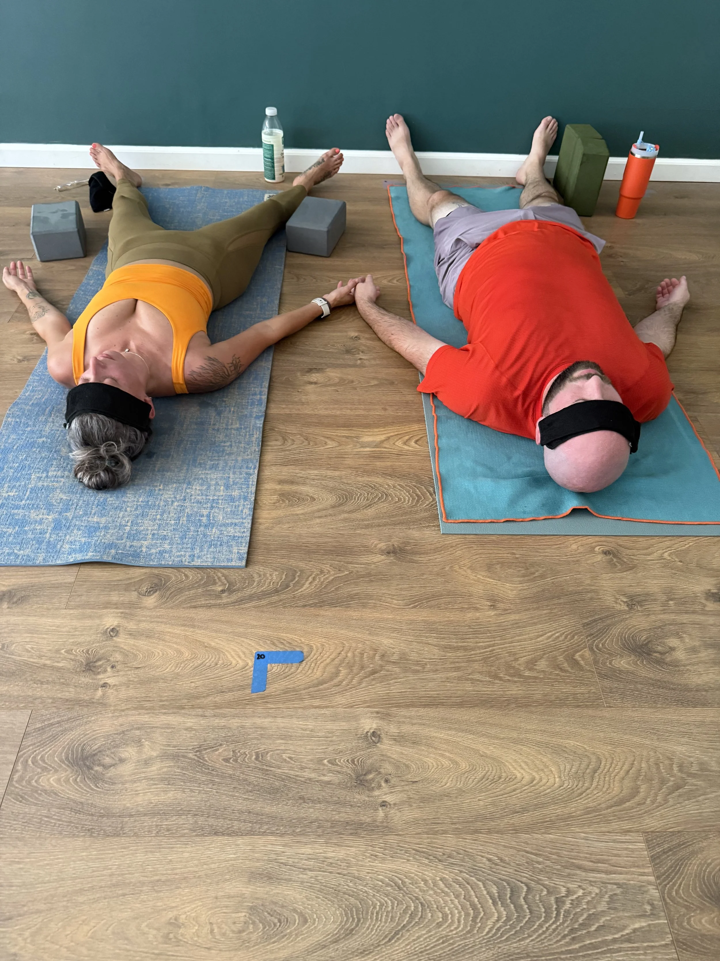 Two people lying on exercise mats doing relaxation or stretching exercises, with yoga blocks, water bottle, and foam roller nearby in a room with wooden floor and teal wall.