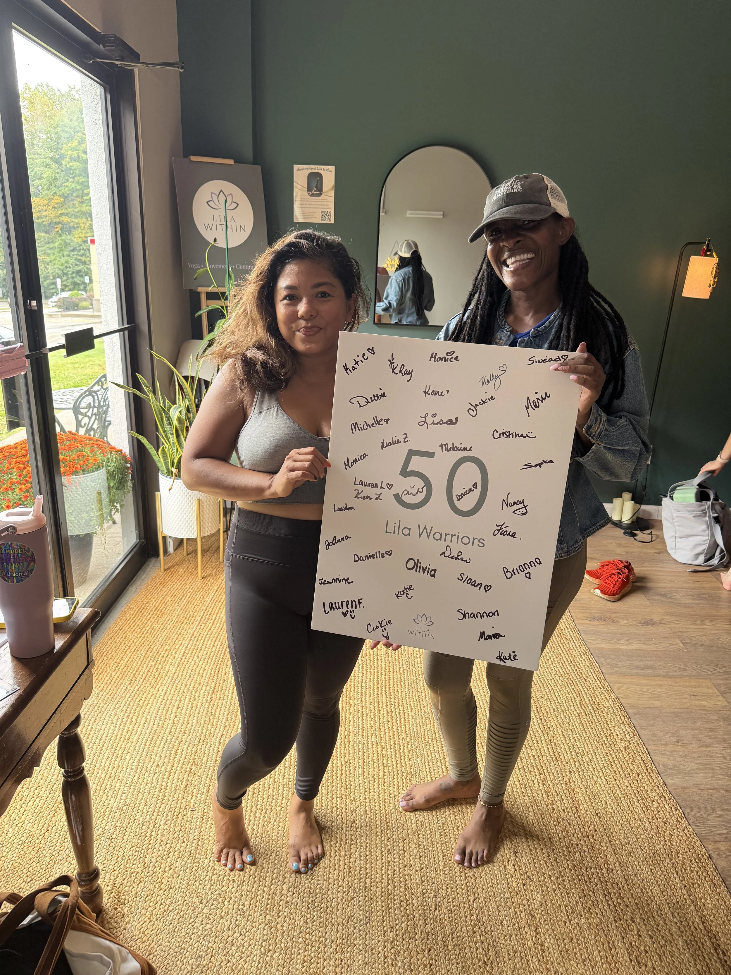Two women standing indoors, holding a large white poster board with handwritten names and a large number 50. They are barefoot on a beige carpet, smiling at the camera. One woman has long dark hair, the other wears a cap and has dreadlocks.