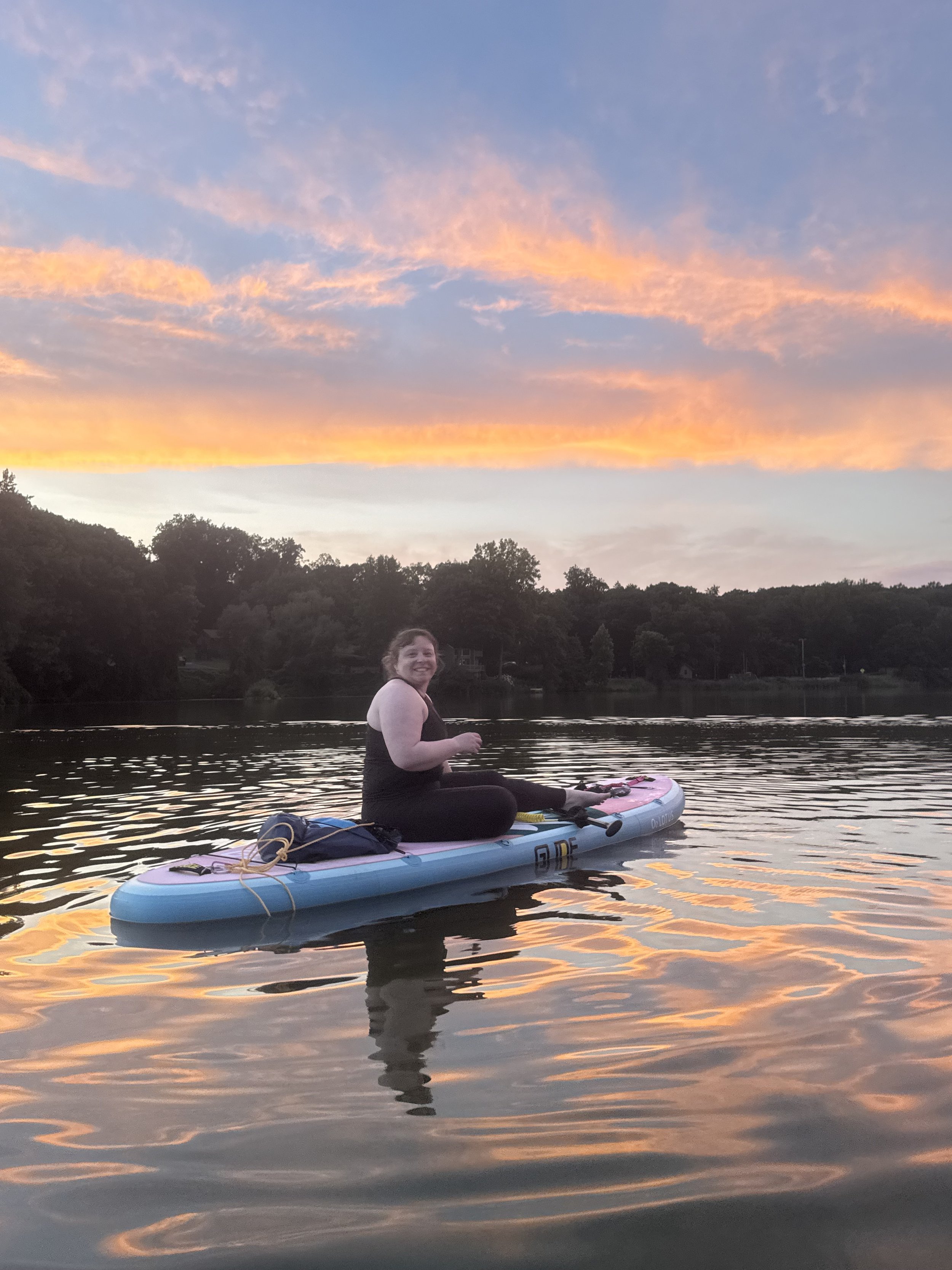 A woman sitting on a paddleboard on a calm lake during sunset, with a colorful sky and distant trees in the background.