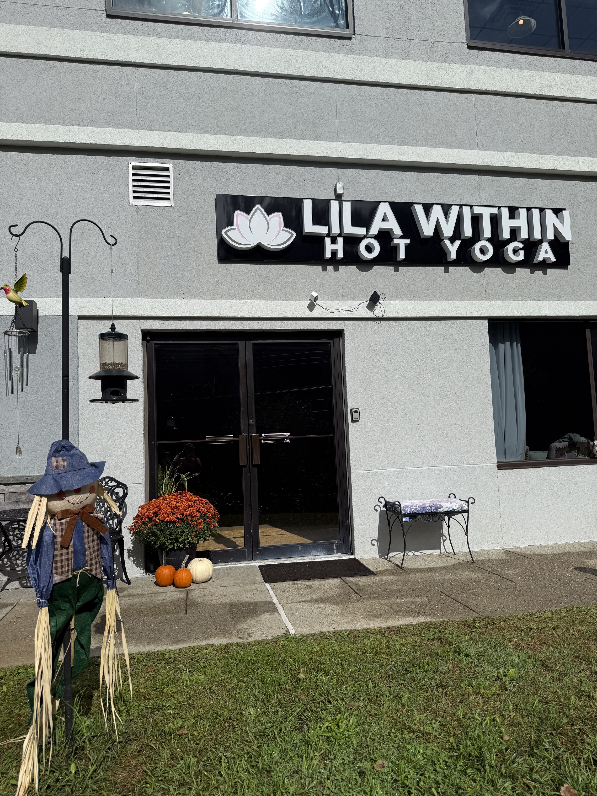 An exterior view of a building with a sign that reads 'Lila Within Hot Yoga' above the entrance. There are pumpkins, a scarecrow decoration, and a potted plant outside the door, indicating a fall or Halloween setting. A black mat is on the ground in 
