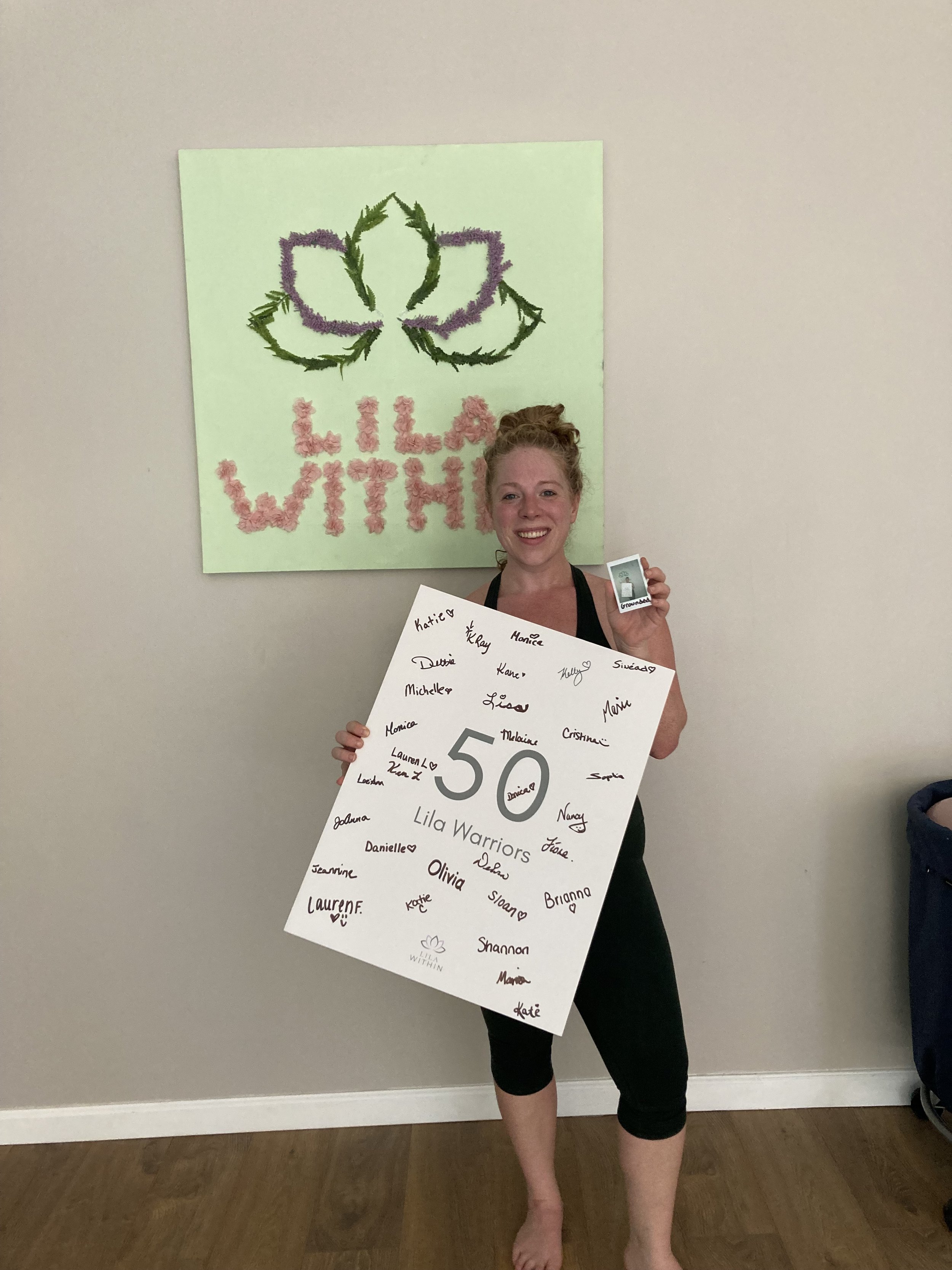 A woman is standing indoors, smiling, holding a large white poster with signatures and the number 50 on it. She is barefoot, wearing black leggings and a black top, standing in front of a wall with a green decorations and a sign reading 'LILA WITHIN'