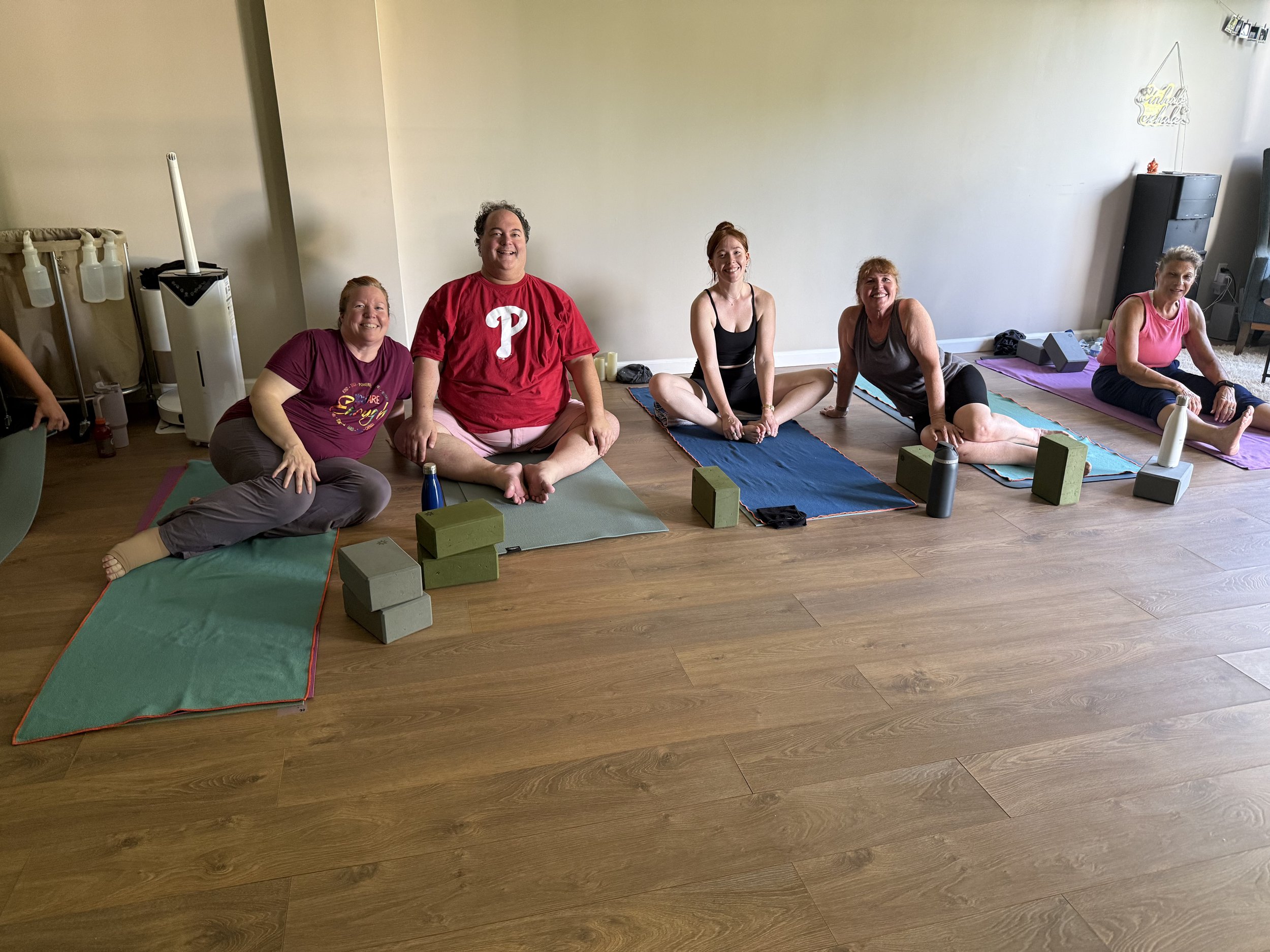 A group of five people sitting on yoga mats, smiling, in a room with hardwood floors, yoga blocks, water bottles, and a plain wall.