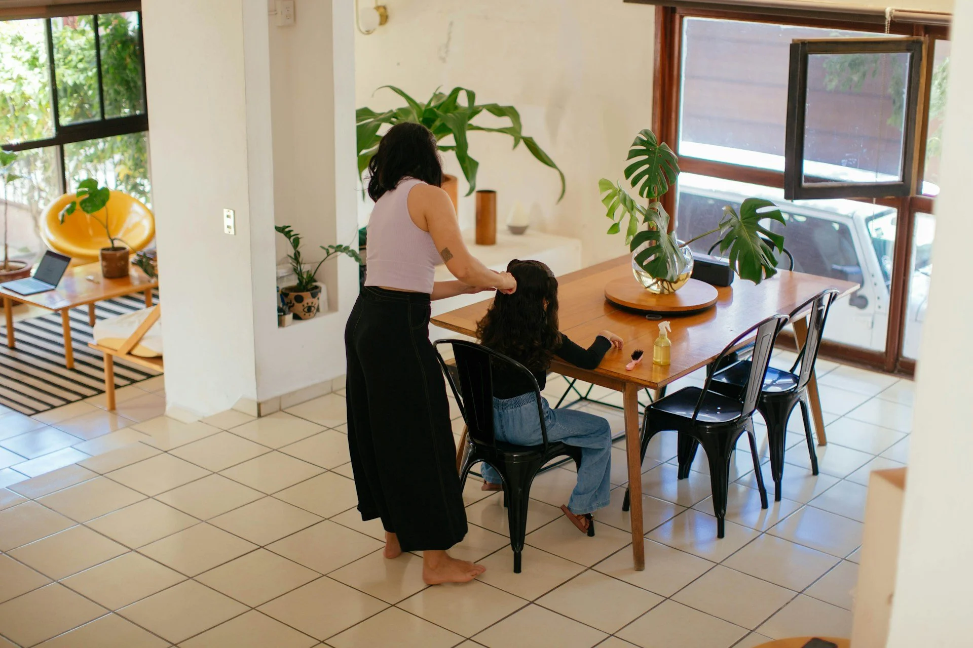 A woman with dark hair and a tattoo on her arm is giving a haircut to a young girl with long dark hair, sitting at a wooden dining table in a bright room with large windows and green plants.