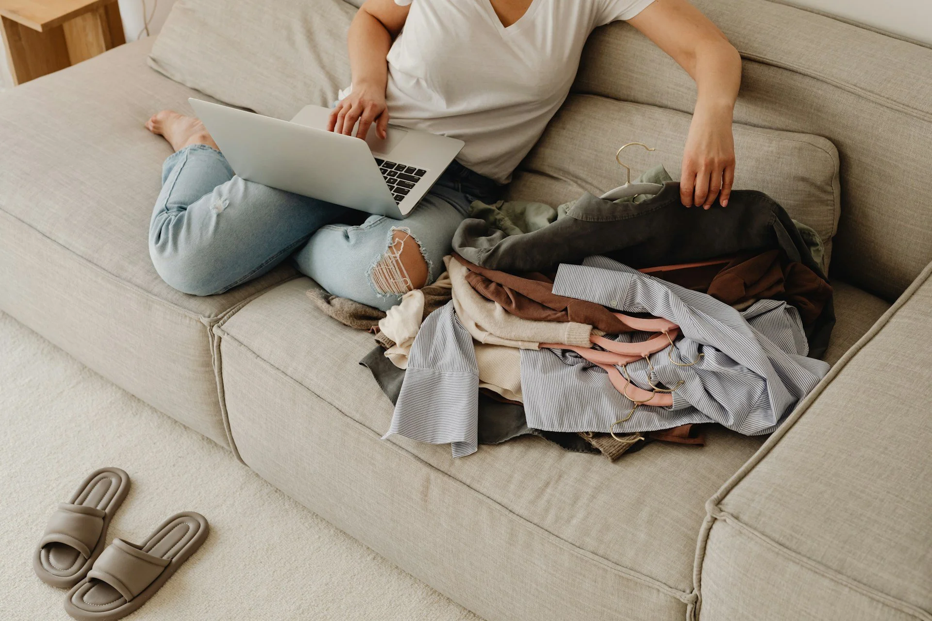 A person sitting on a beige sofa with a laptop, surrounded by scattered clothes, and a pair of beige slippers on a light carpet.