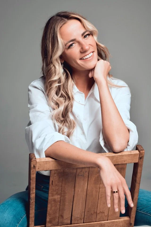 Smiling woman with blonde wavy hair wearing a white shirt, sitting on a chair, against a neutral background.