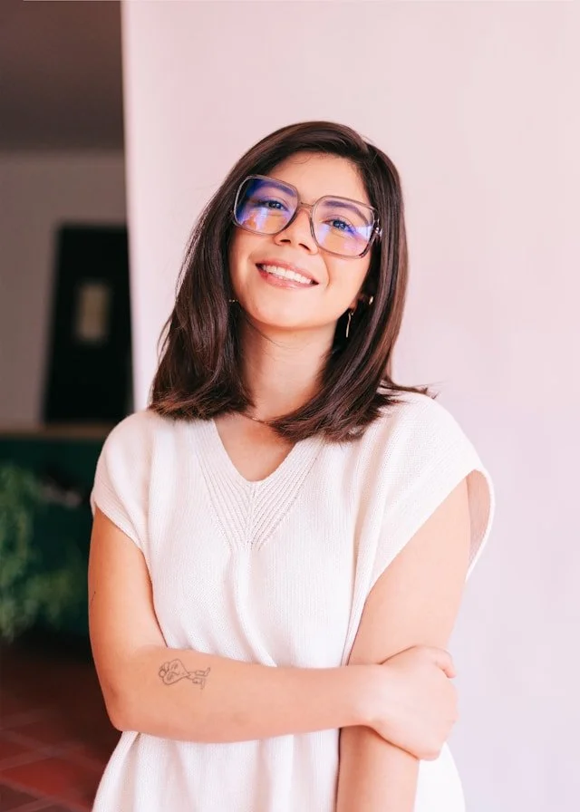 Smiling woman with shoulder-length dark hair, wearing glasses, a white sleeveless top, crossing her arms, standing indoors against a pink and white background.