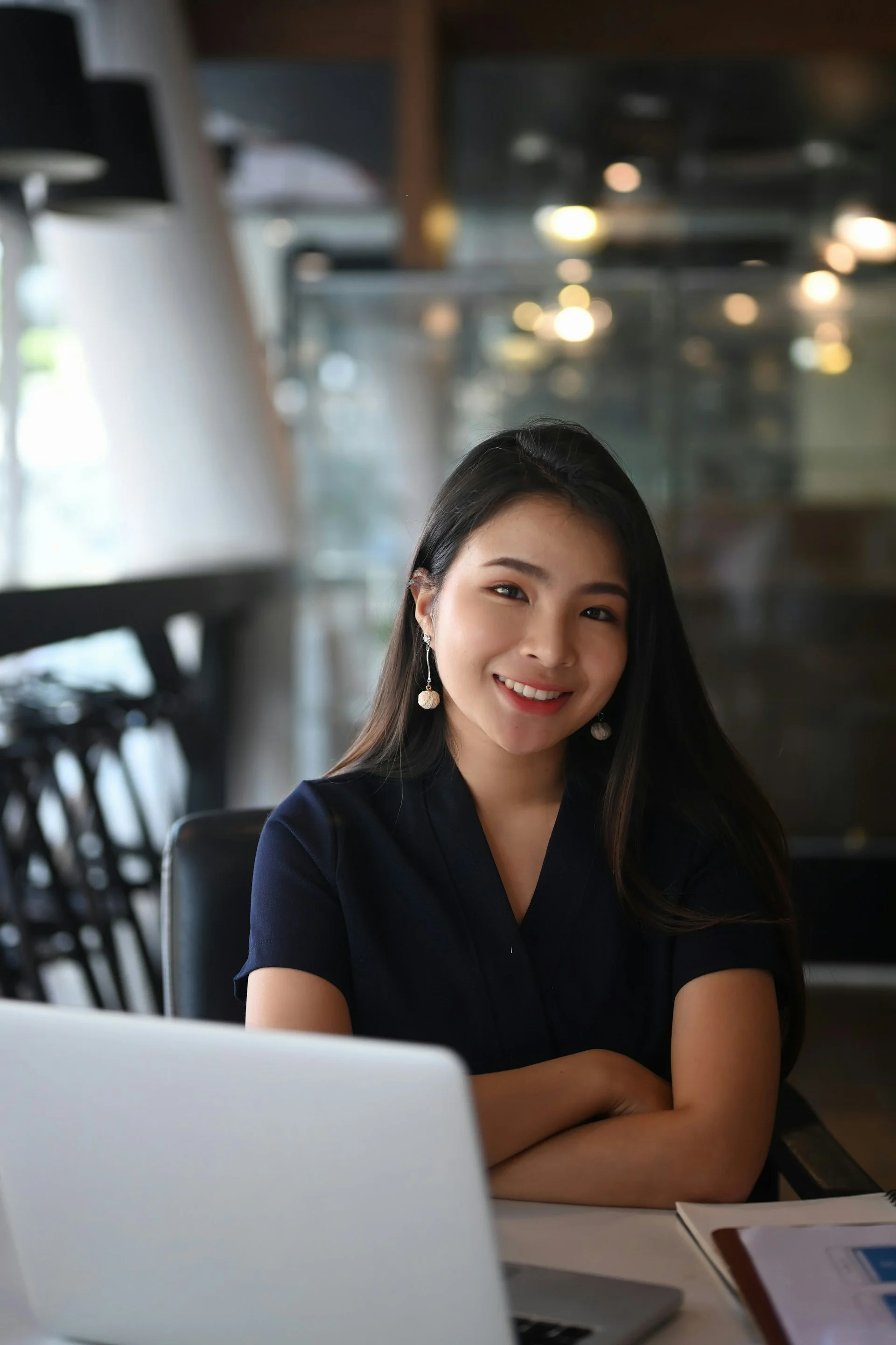 A young woman with long dark hair and earrings smiling while sitting at a table in a modern indoor setting, possibly a cafe or office, with a blurred background.
