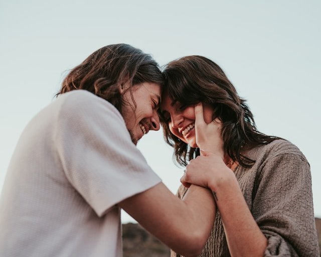 A couple smiling and laughing with their foreheads touching and hands clasped together outdoors.