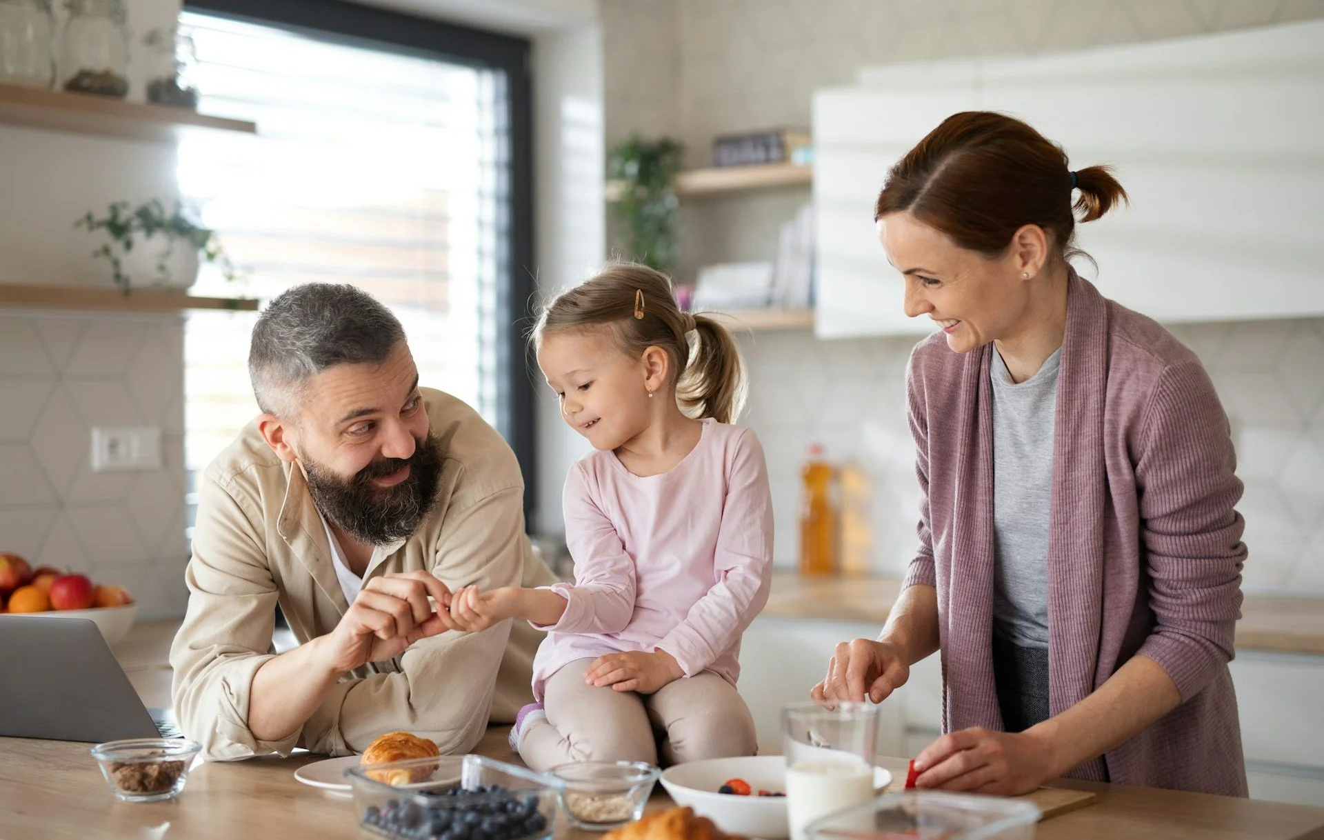A family of three, including a man, a woman, and a young girl, enjoying time together in a bright modern kitchen. They are smiling and interacting near a table with breakfast foods and bowls of fruit, with natural light coming in through a large window.