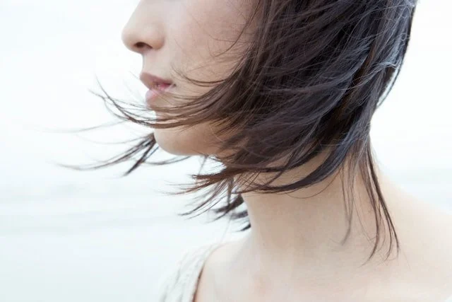 Close-up of a woman's face and neck with windswept short dark hair, light skin, and neutral expression.