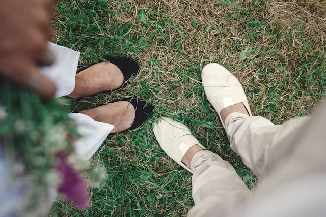 Two people standing on grass, one wearing black shoes and white dress, the other wearing off-white slides and beige pants.