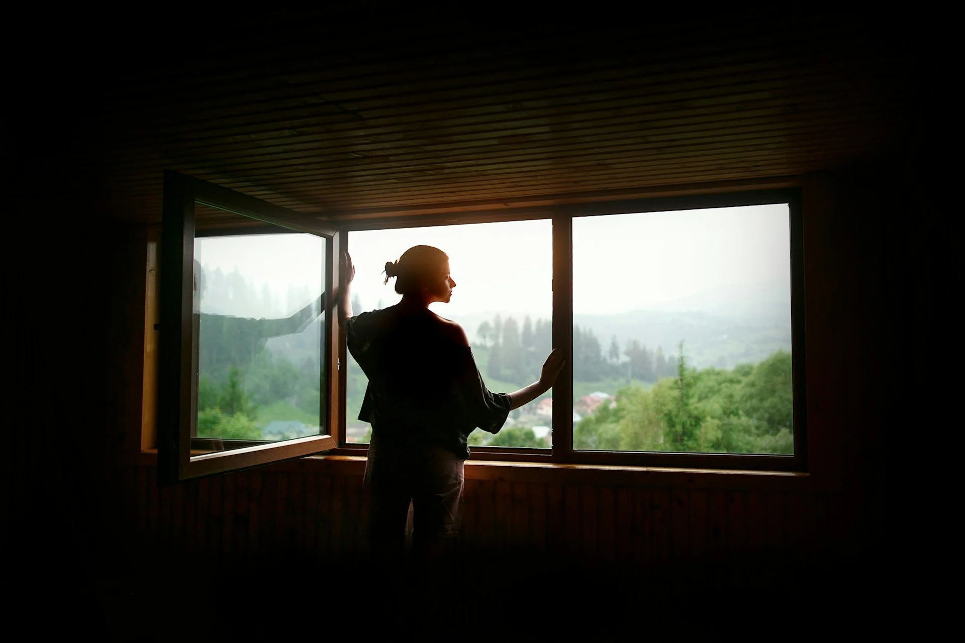 A woman standing in front of a large window, looking out at a landscape with trees and hills during daytime, with her hand on the window frame and the other on the glass.
