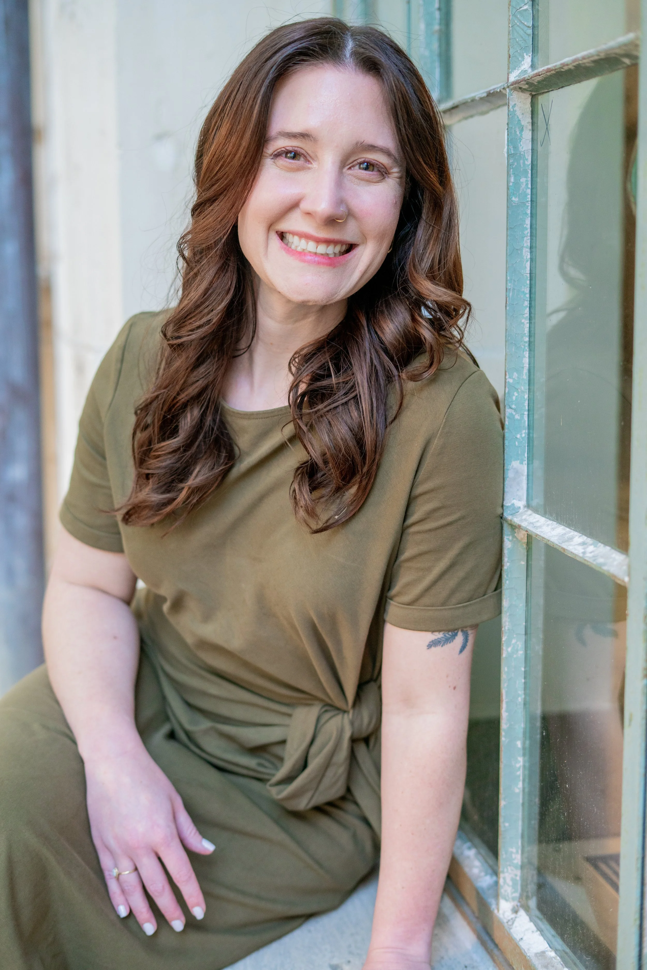 A woman with long brown hair, light skin, and a nose piercing is sitting near a window, smiling at the camera. She is wearing an olive green shirt and pants, with a tattoo visible on her forearm. Her left hand rests on her knee, showing a ring on her finger.