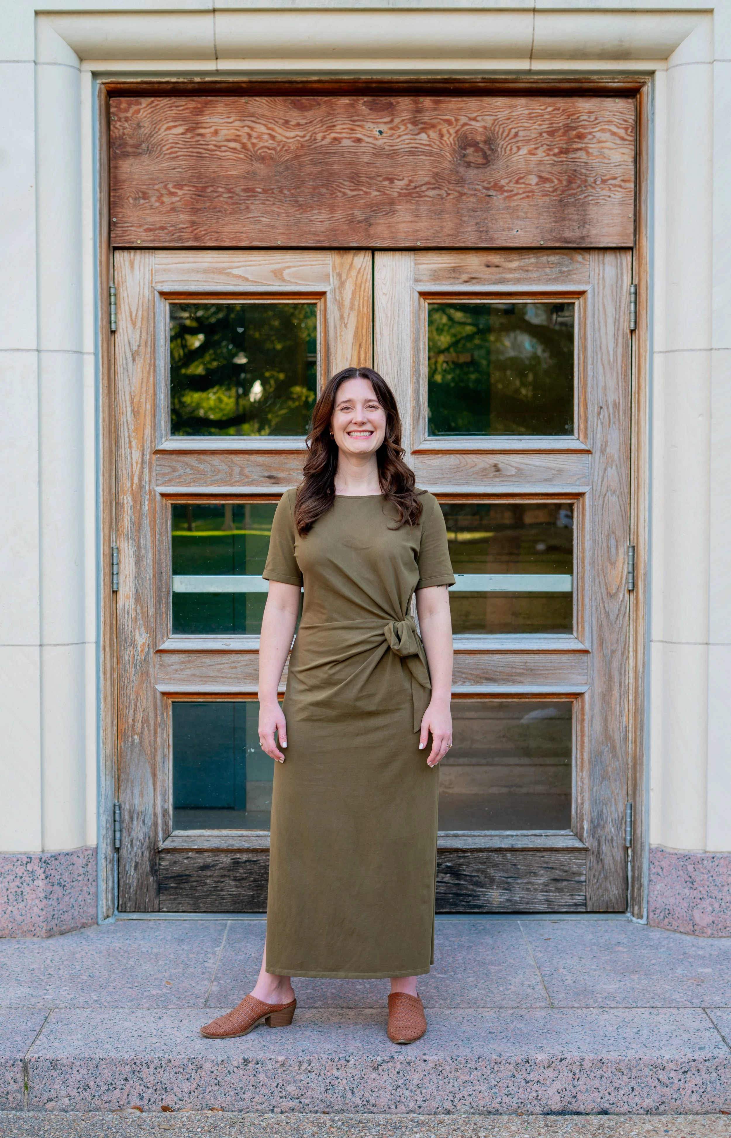 A woman with long brown hair smiling, standing in front of a wooden and glass door, wearing a green dress and brown shoes.