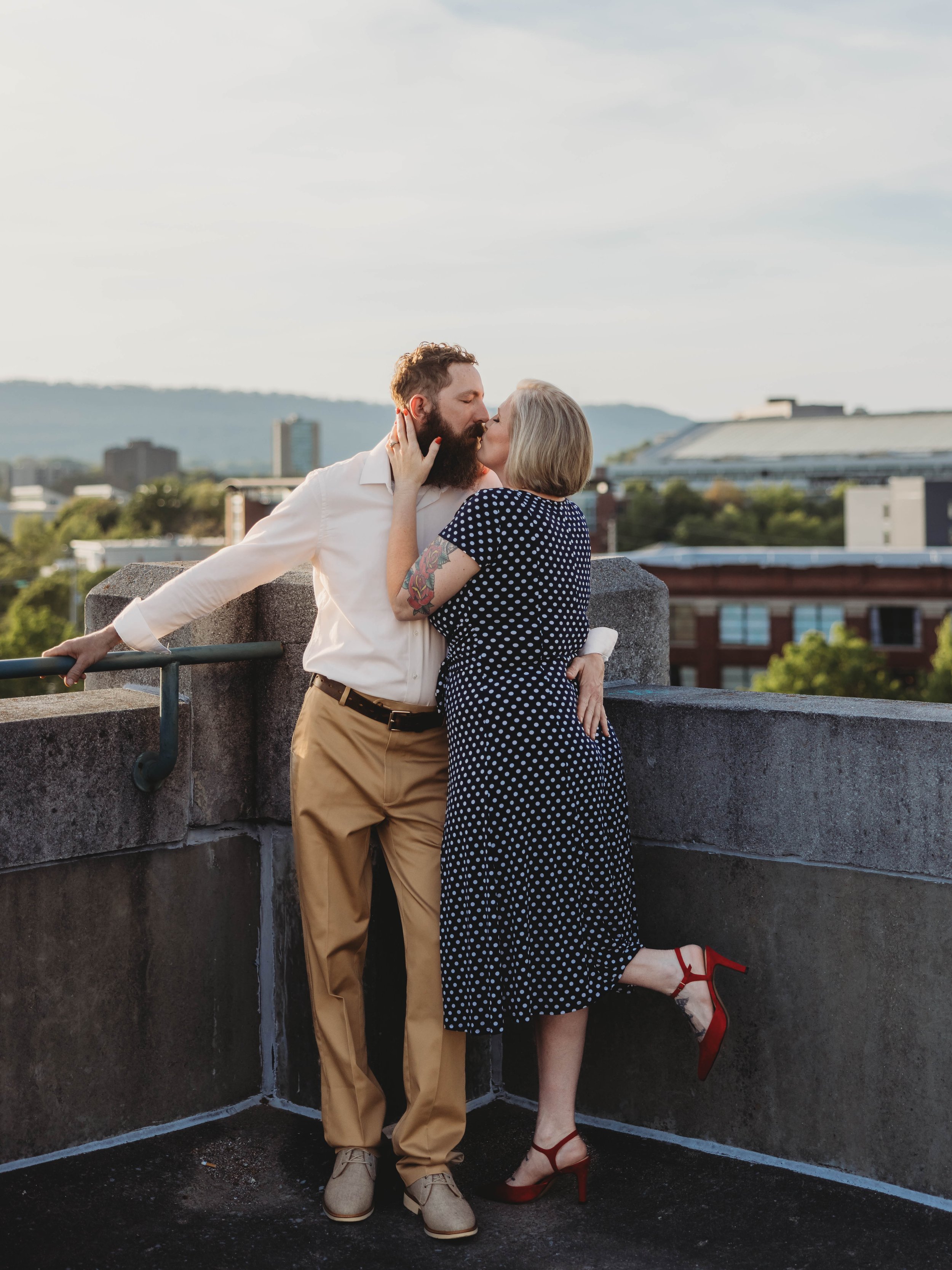 A man with a beard and a woman with blonde hair are sharing a kiss on a rooftop. The man is wearing a white shirt and khaki pants, while the woman is dressed in a navy polka-dot dress and red high heels. The city skyline is visible in the background.