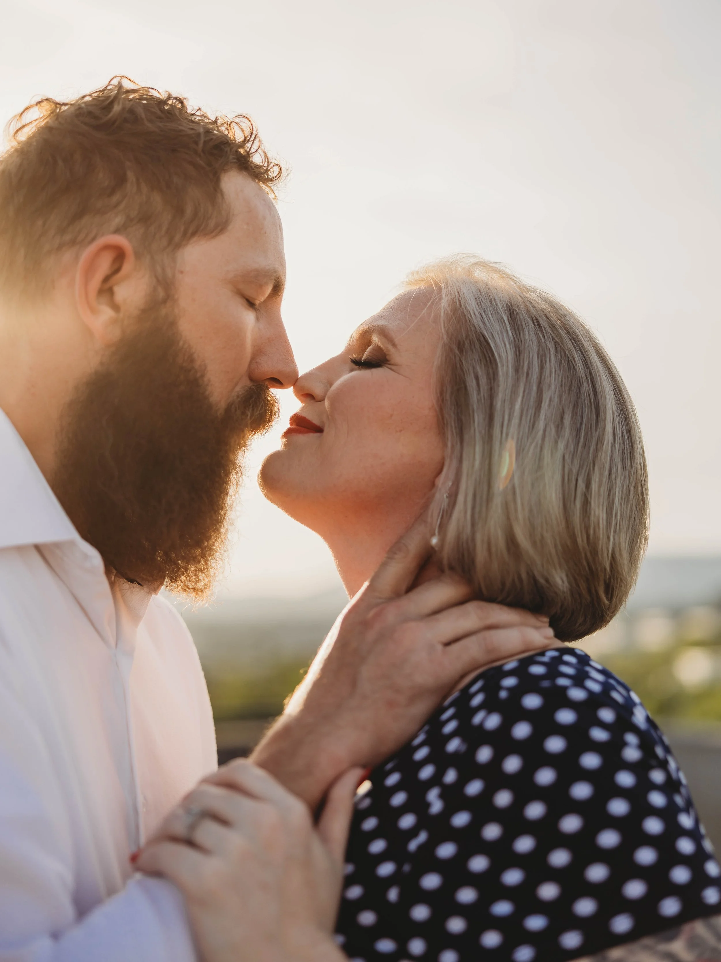 A young man with a beard and closed eyes gently touching the face of an elderly woman with gray hair and closed eyes, both with peaceful expressions, outdoors during sunset.