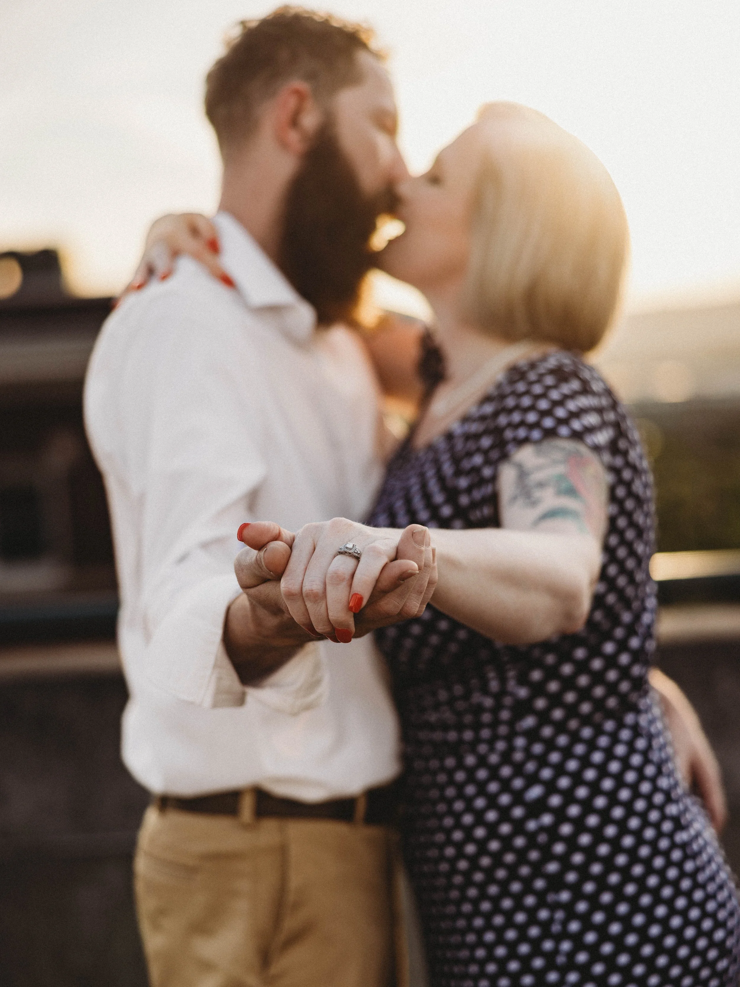 A man and woman are kissing while holding hands outdoors during sunset.