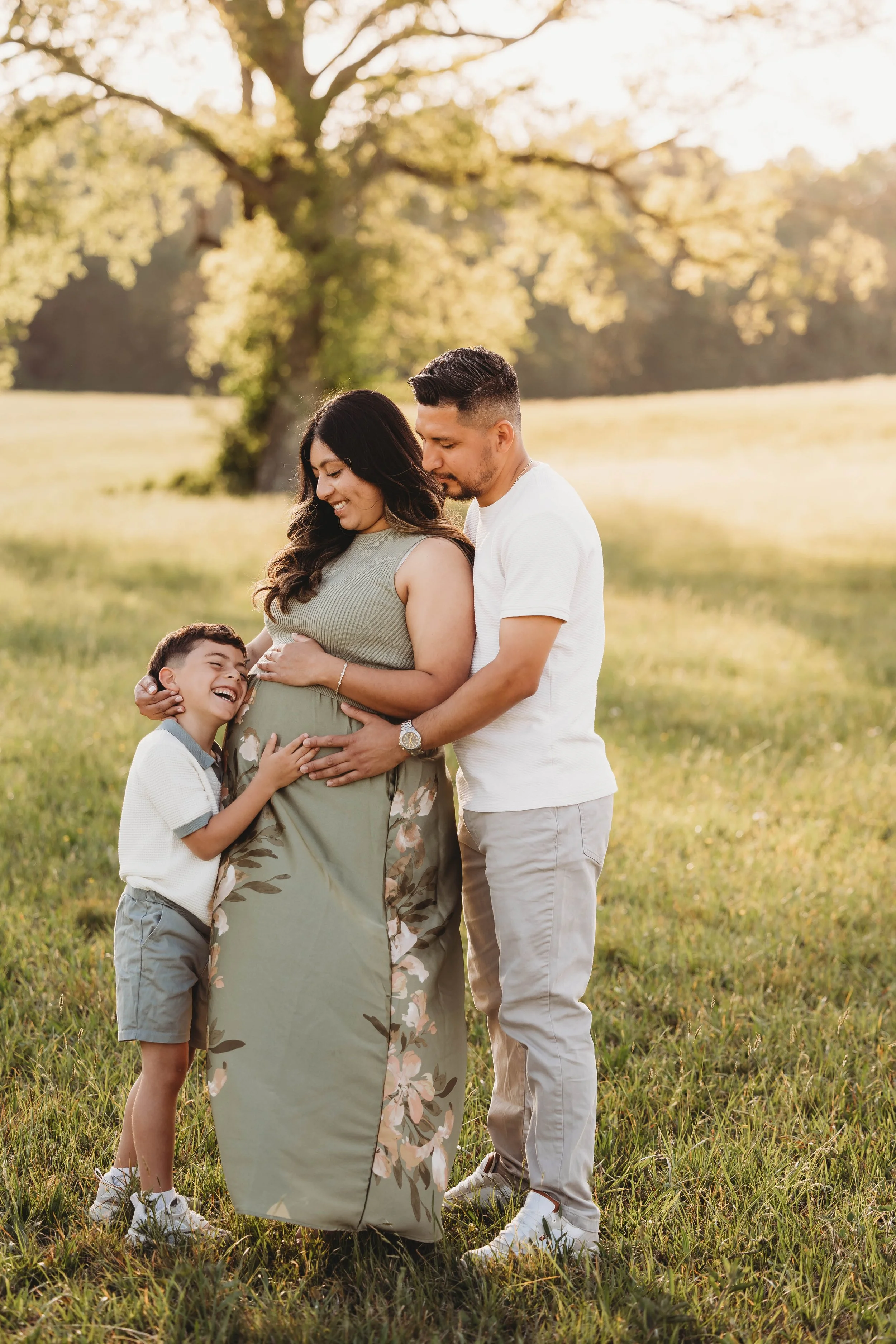 A happy family of four, including a pregnant woman, a man, and two children, enjoying a moment outdoors in a grassy field with trees in the background during sunset.