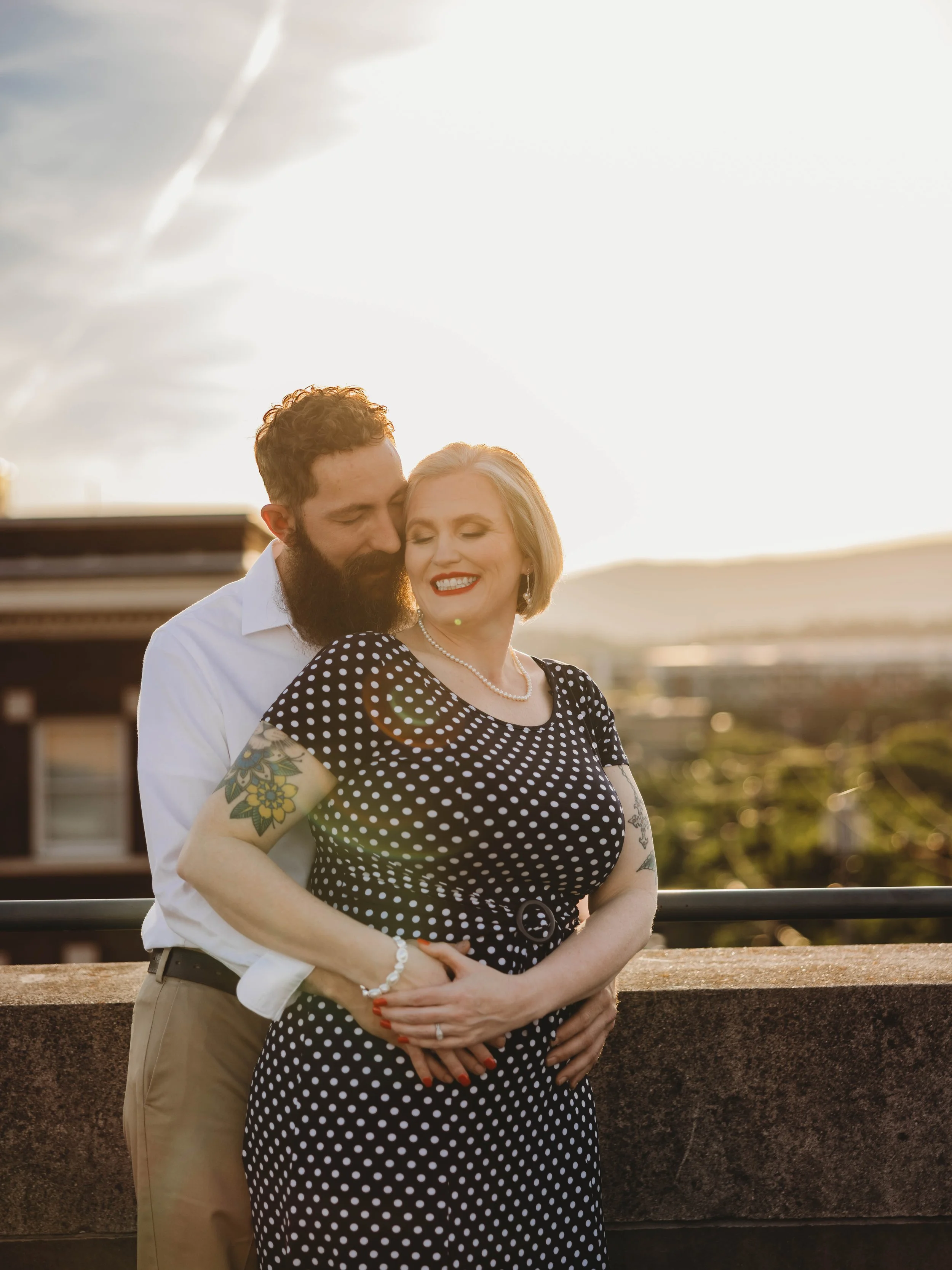 A man and woman embrace outdoors during sunset, with the man kissing the woman's temple. The woman is smiling with eyes closed, wearing a black polka dot dress, pearl necklace, earrings, and tattoos on her arms. The man has a beard, mustache, and sho