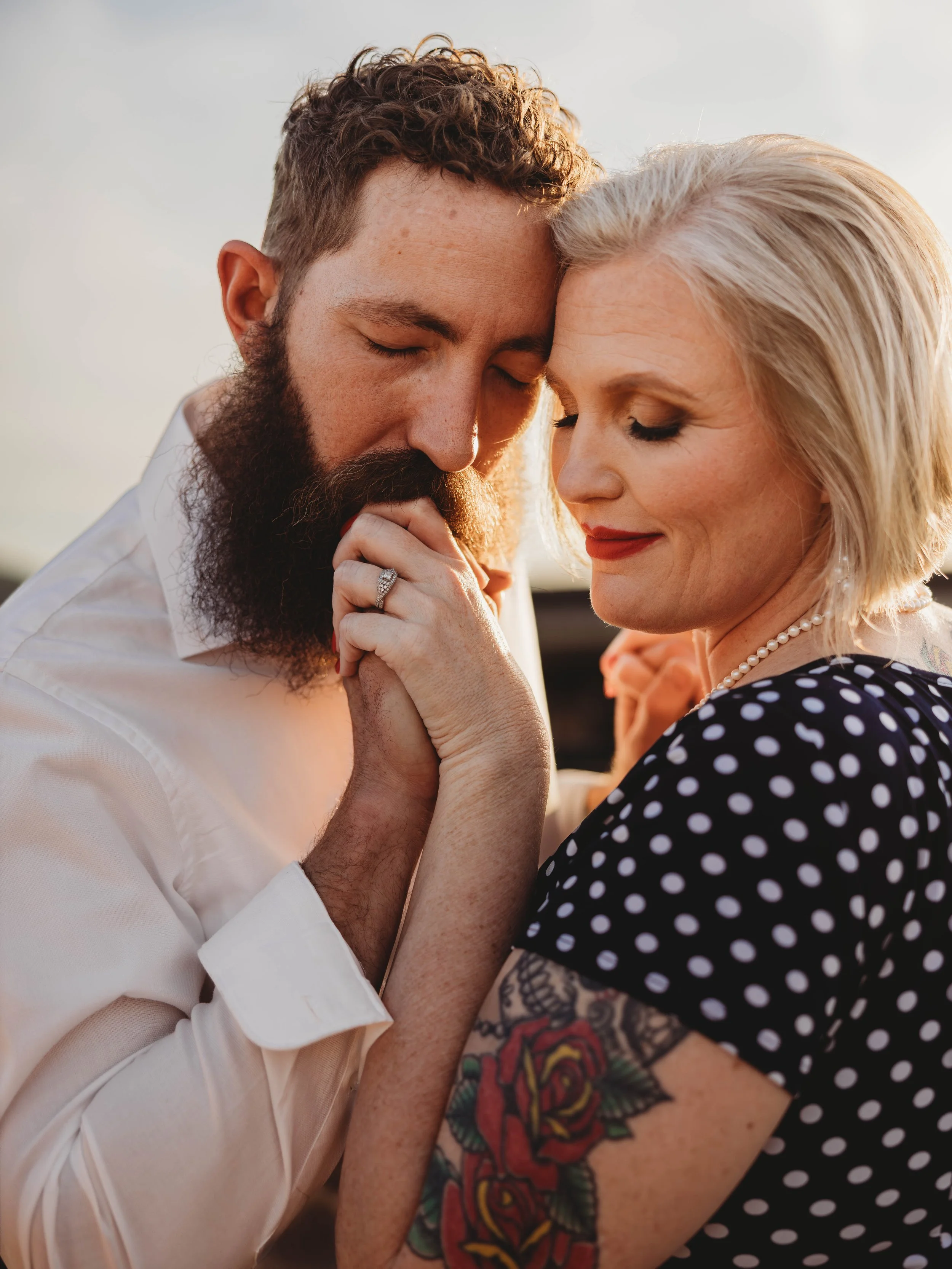 A man with a beard and a woman with blonde hair hold their foreheads and noses together, eyes closed, during a tender moment outdoors in warm sunlight.