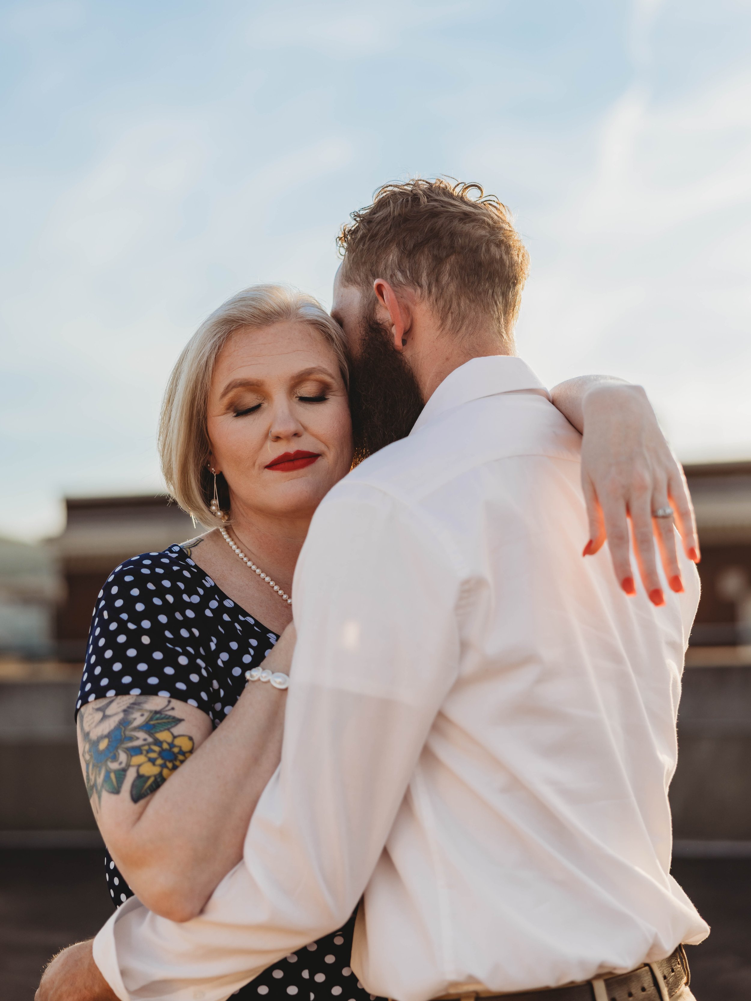 A couple hugging outdoors with the woman closing her eyes and smiling, around sunset or evening time.