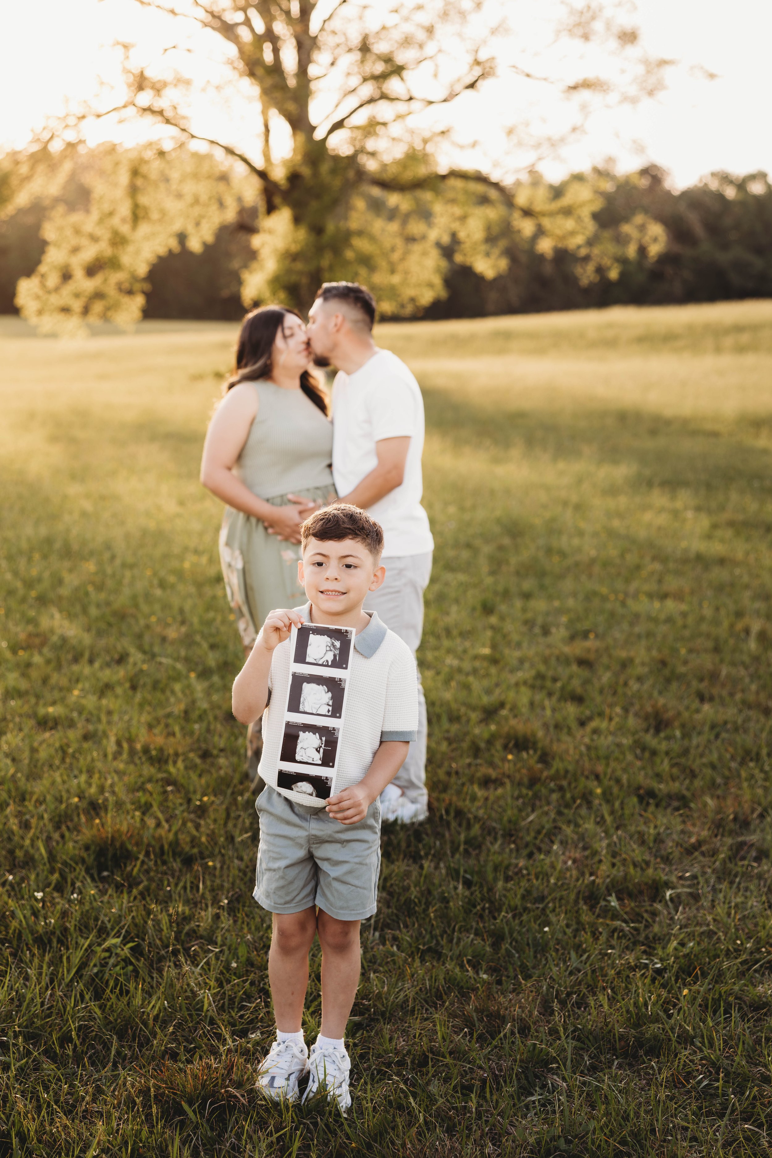 Young boy holding ultrasound images in front of a grassy field during a sunset, with a couple embracing and kissing in the background, under a large tree.