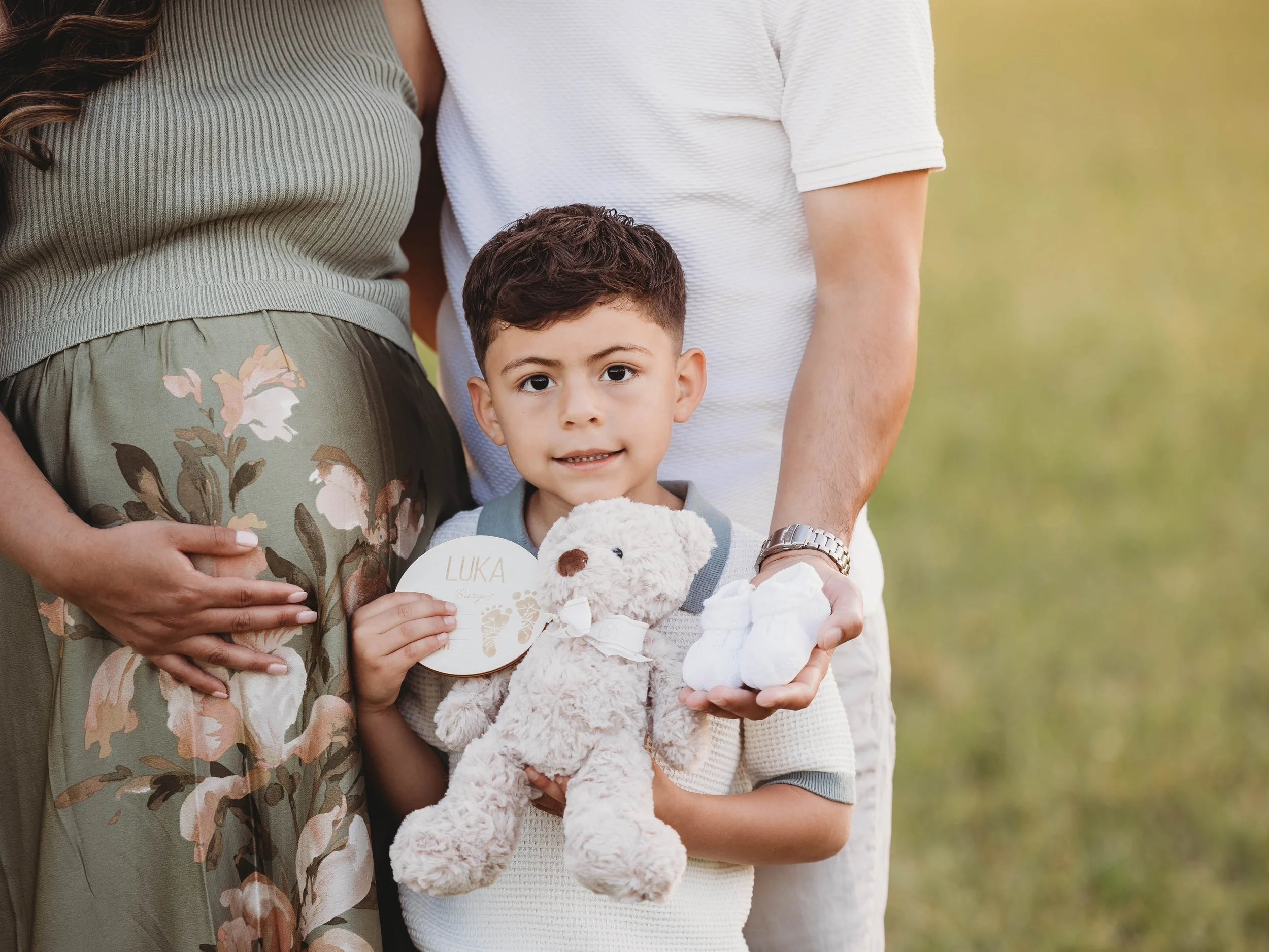 Close-up of a young boy holding a teddy bear, standing between two adults outdoors on a grassy background.