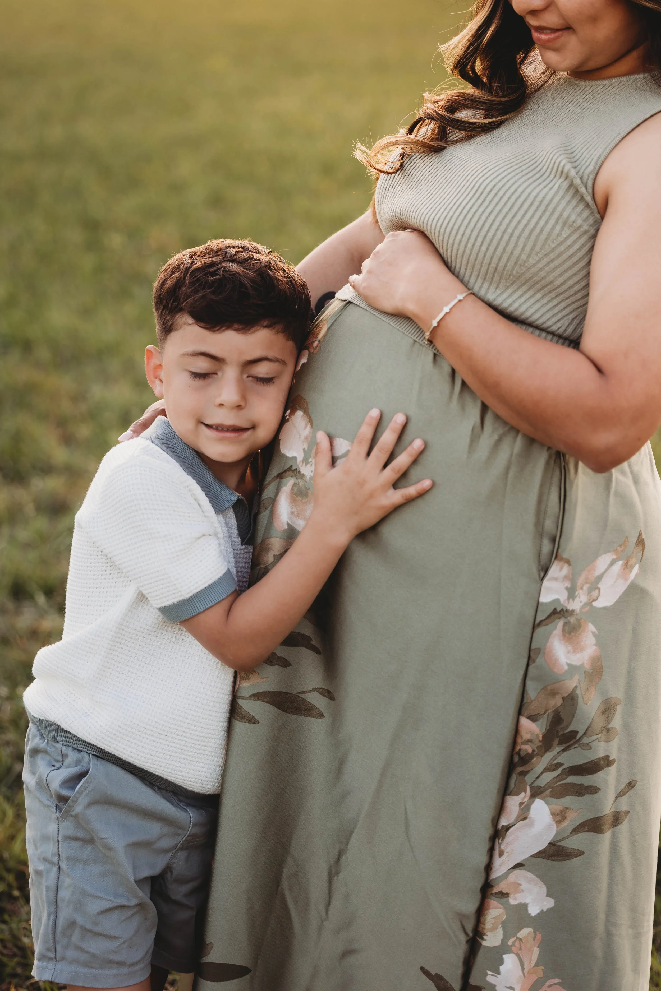 A pregnant woman in a beige sleeveless top and a floral skirt gently holding her belly, with a young boy hugging her from the side, both with their eyes closed and smiling in an outdoor setting.