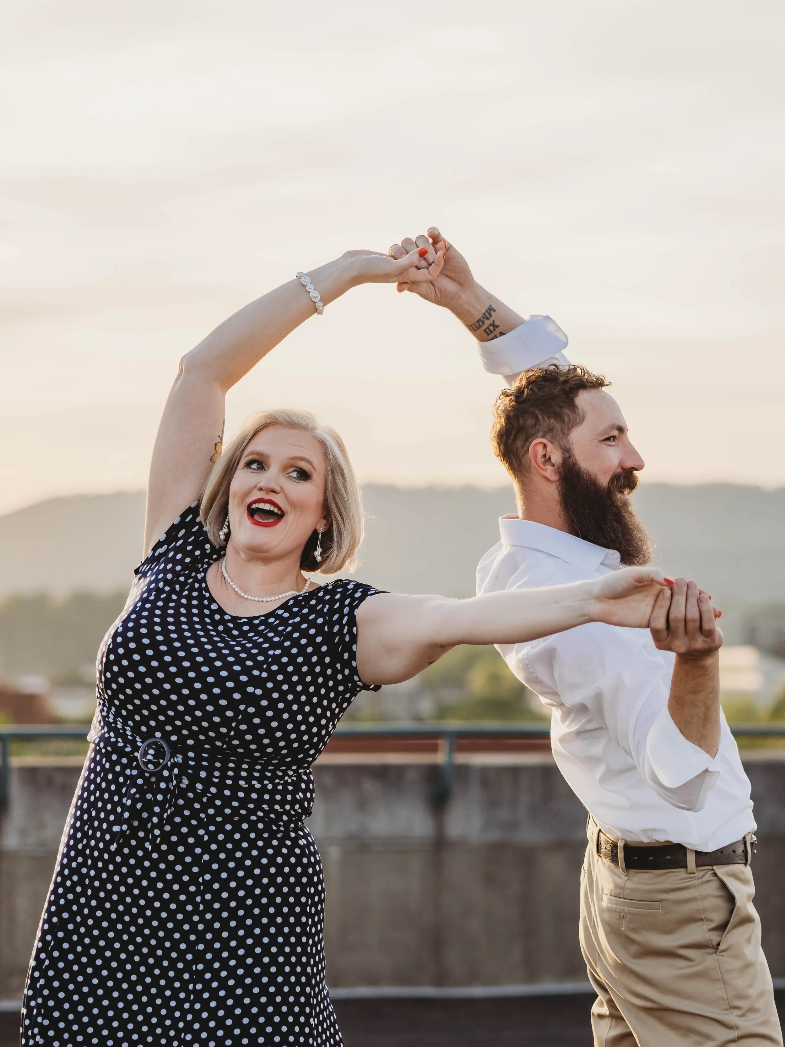 A woman and a man are dancing together outdoors during sunset, holding hands and smiling, with scenic hills in the background.